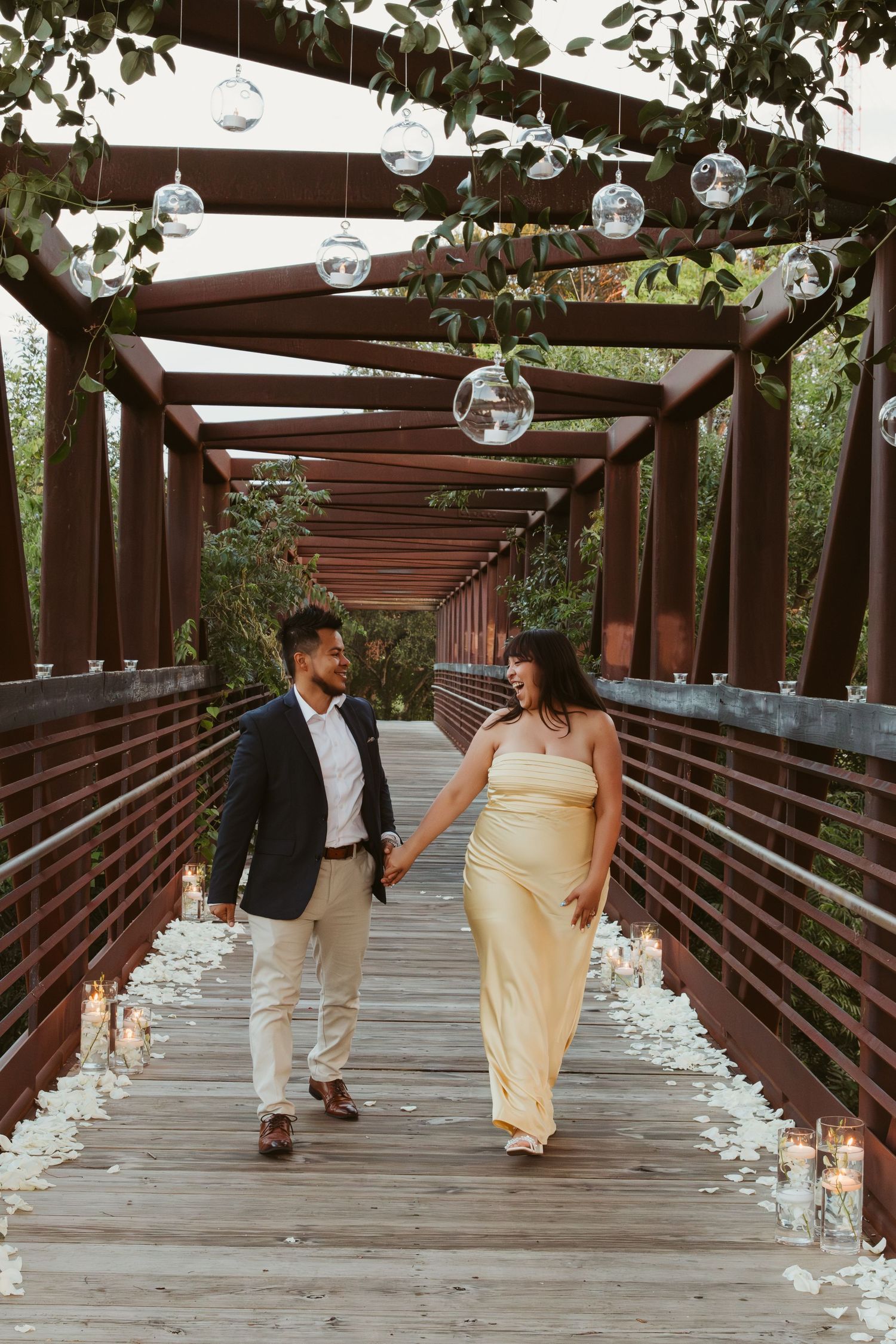 A couple walks along a wooden bridge walkway decorated with hanging glass globes and candles on the ground.