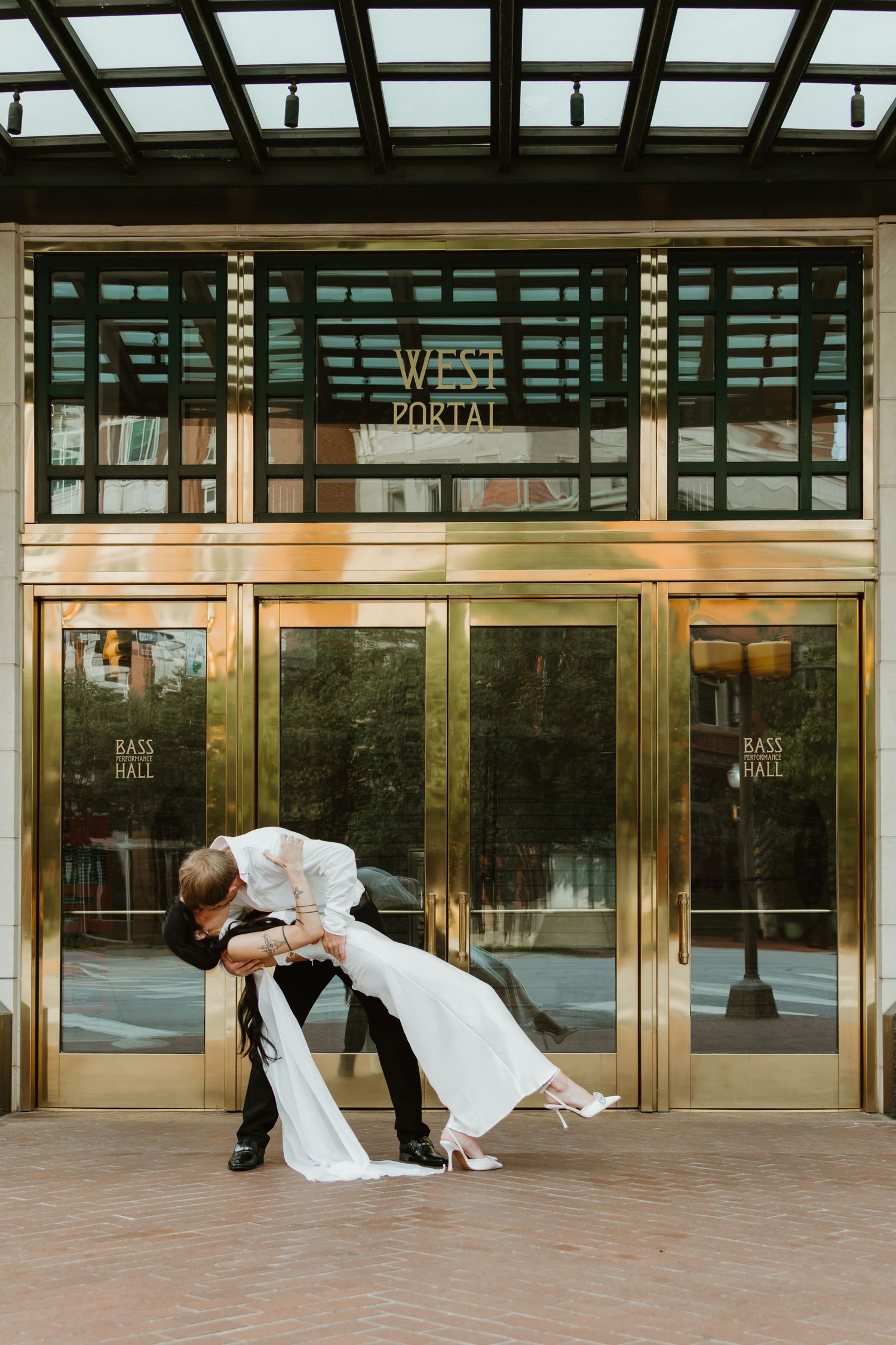 A couple shares a romantic dip kiss pose in front of ornate glass doors at the entrance of a hotel or office building.