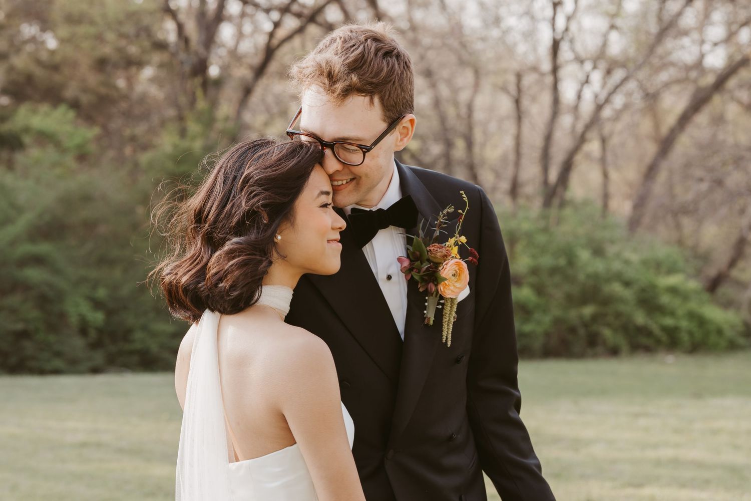 A bride and groom share a romantic moment outdoors, with the groom in a black tuxedo and boutonniere nuzzling the smiling bride.