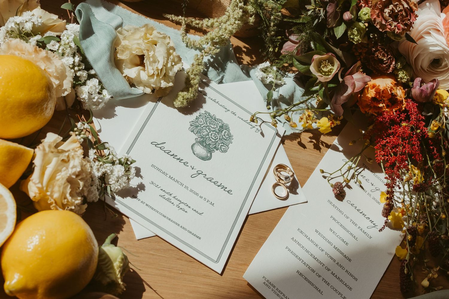 Elegant wedding stationery flat lay with lemons, florals, and gold rings on a wooden surface.