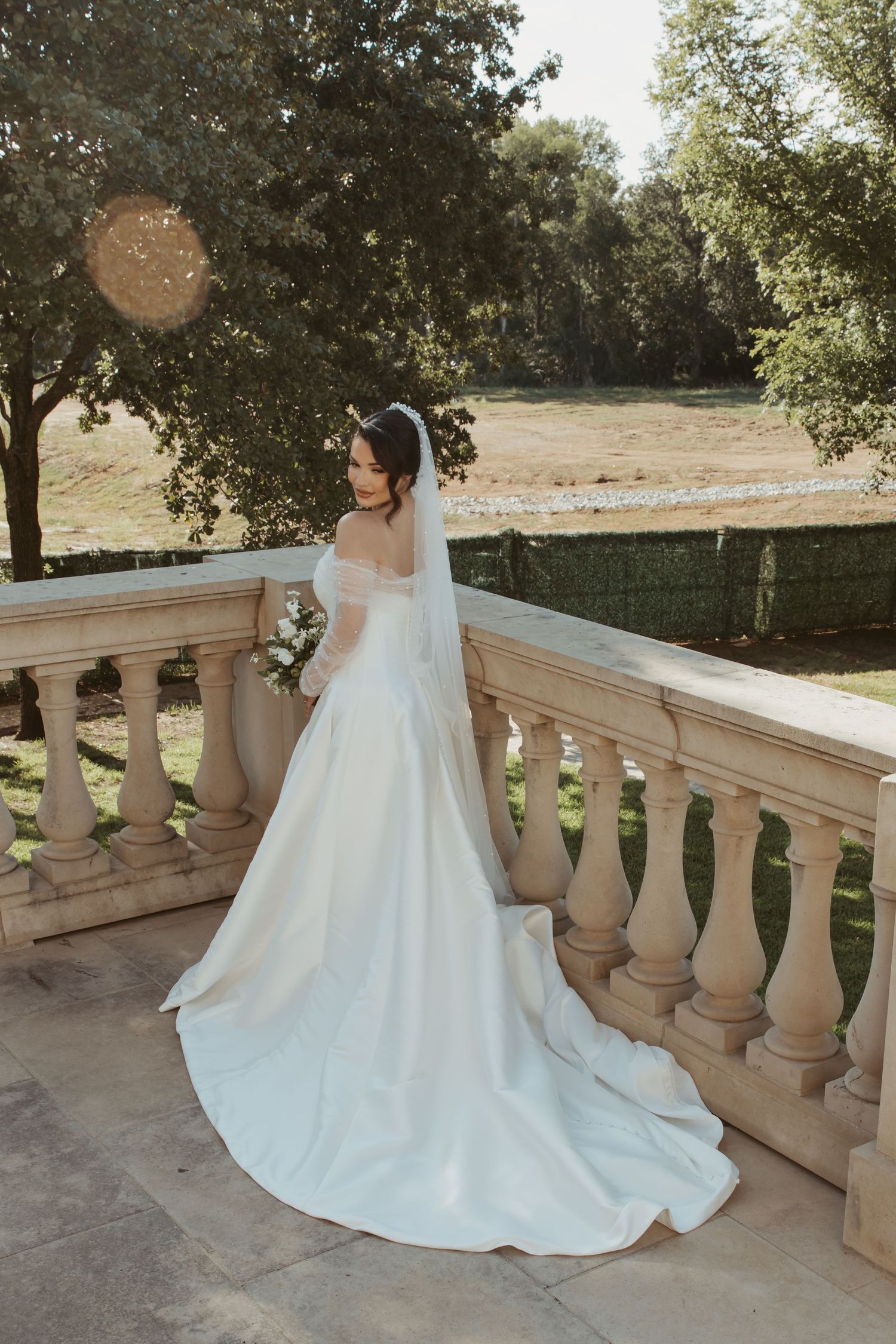 A bride in a flowing white wedding gown with a long train poses on a stone balcony overlooking a sunlit garden.
