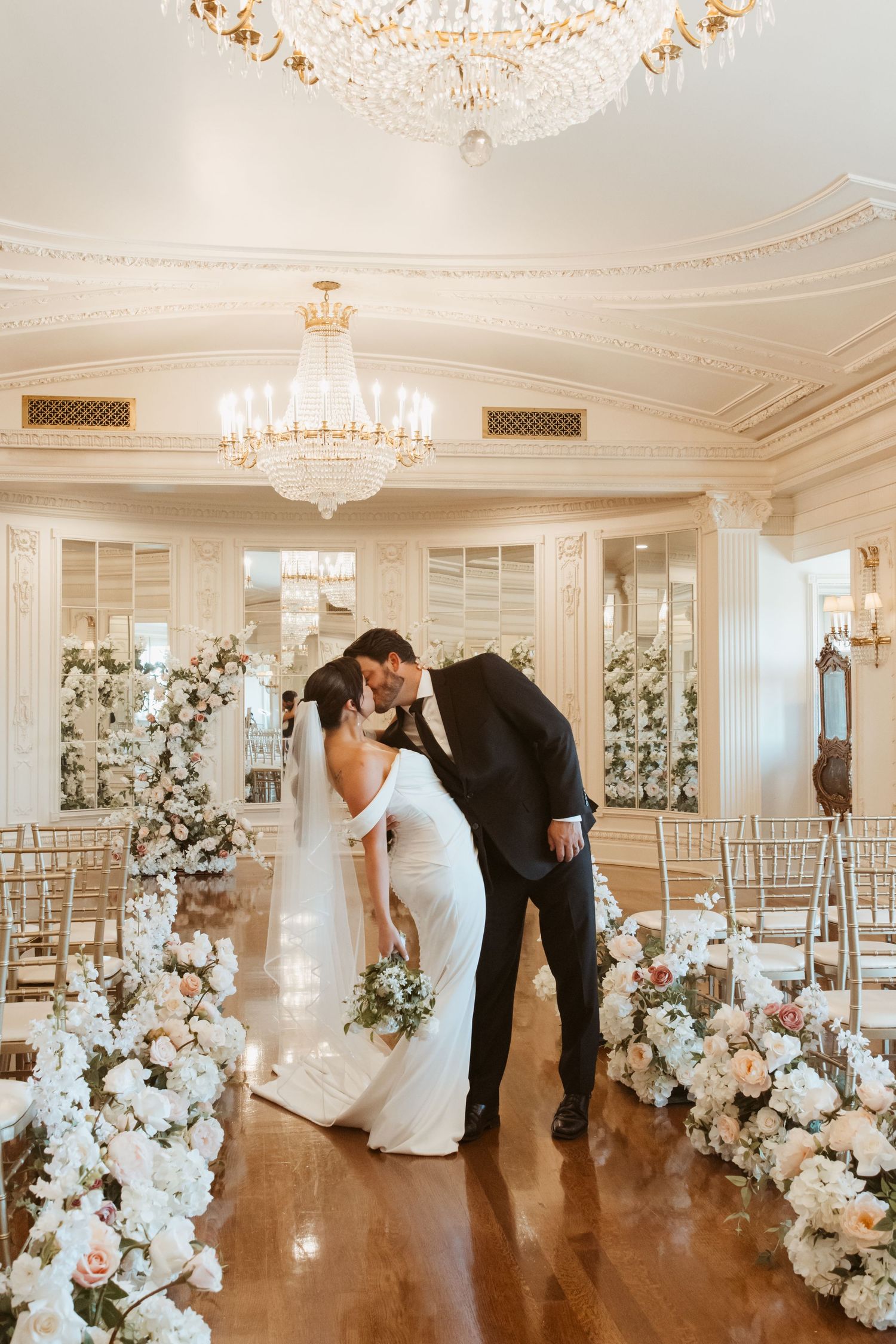 Elegant white and gold ballroom decorated with luxurious floral arrangements and crystal chandeliers for a wedding celebration.
