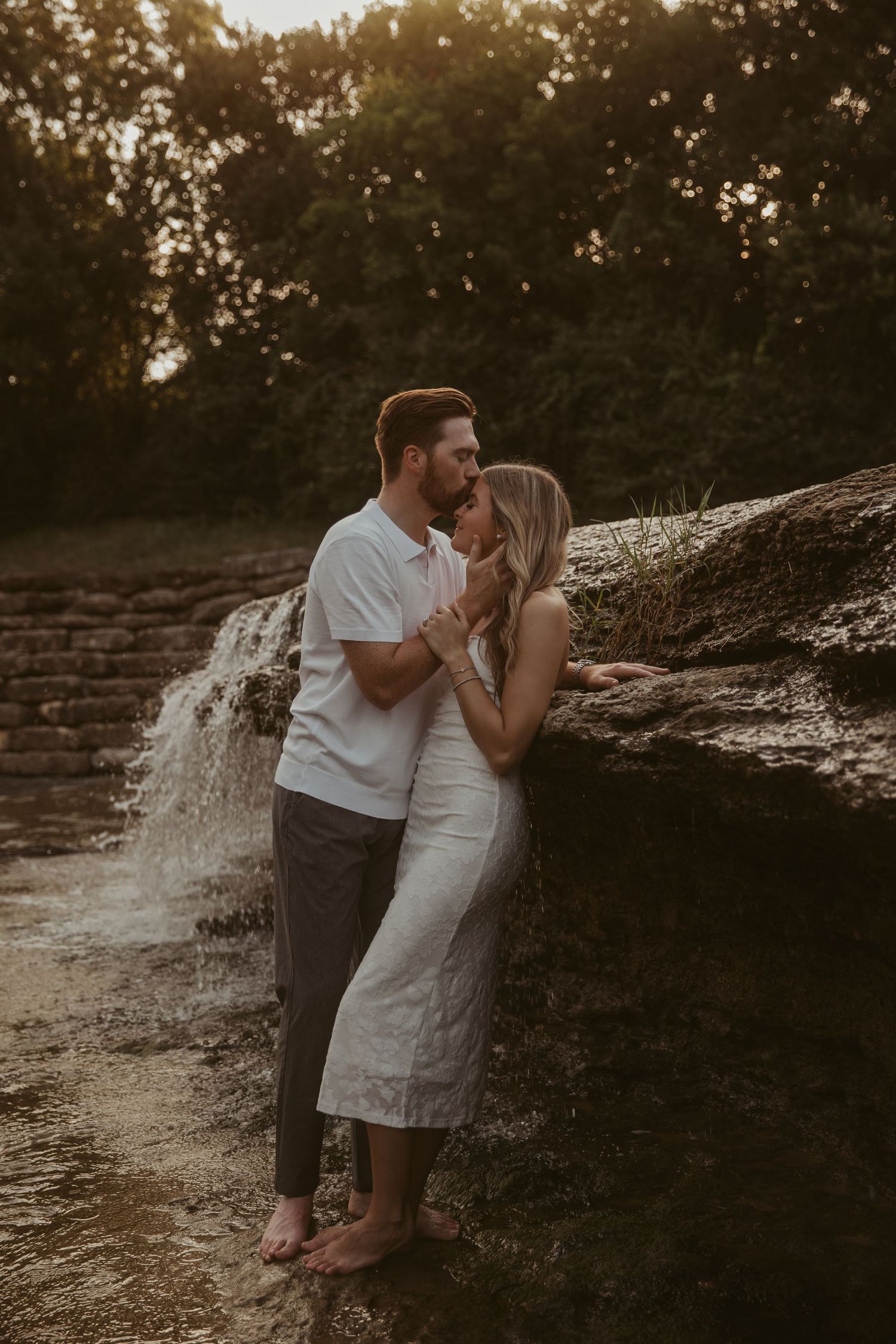 Romantic couple sharing a kiss by a waterfall, woman in white dress, man in white shirt, golden hour lighting.