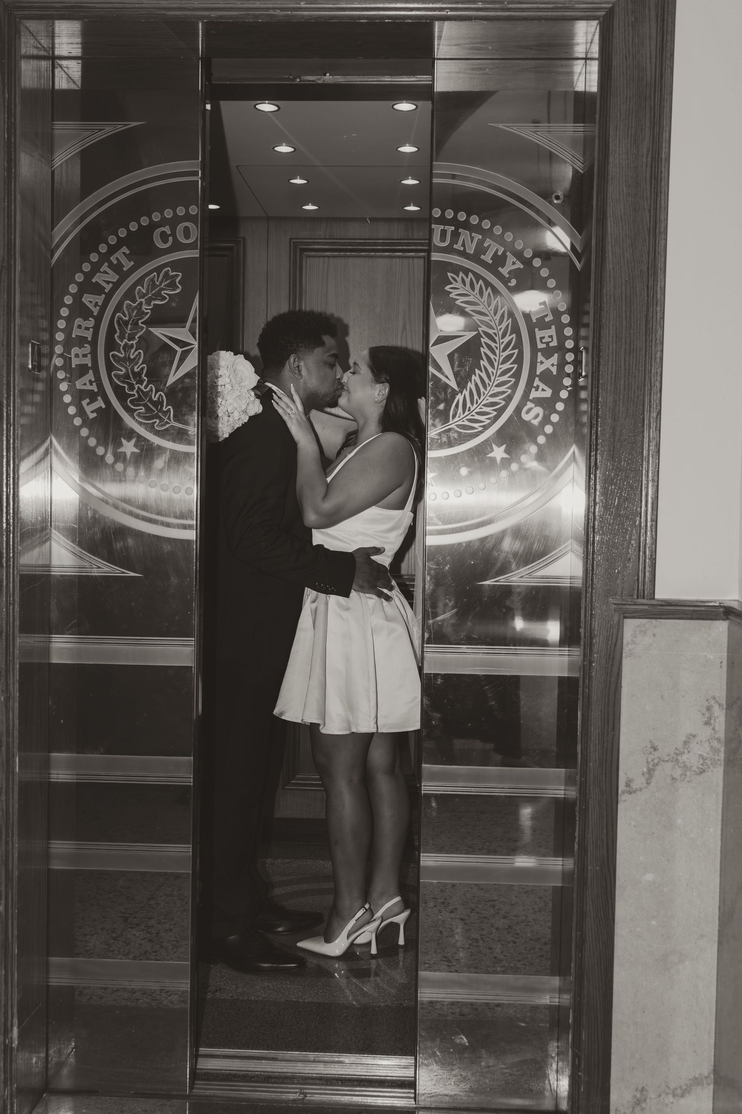A couple shares a kiss in front of reflective glass doors with decorative emblems. Wedding Photographer DFW