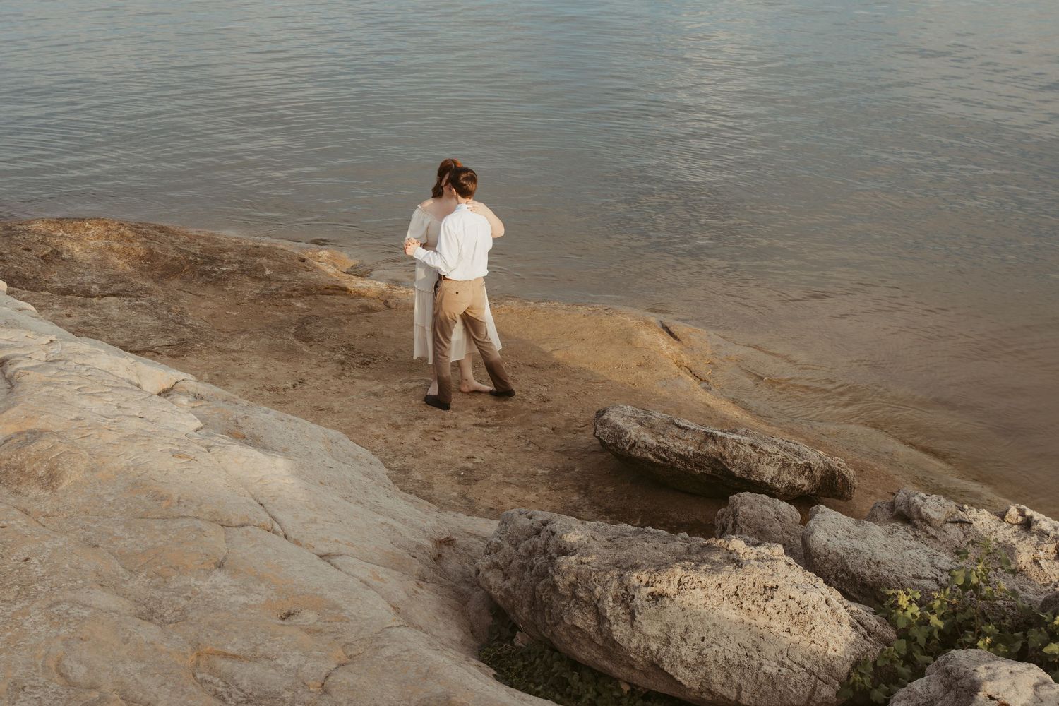 A couple in light colored clothing standing together on rocky coastal terrain by calm waters.
