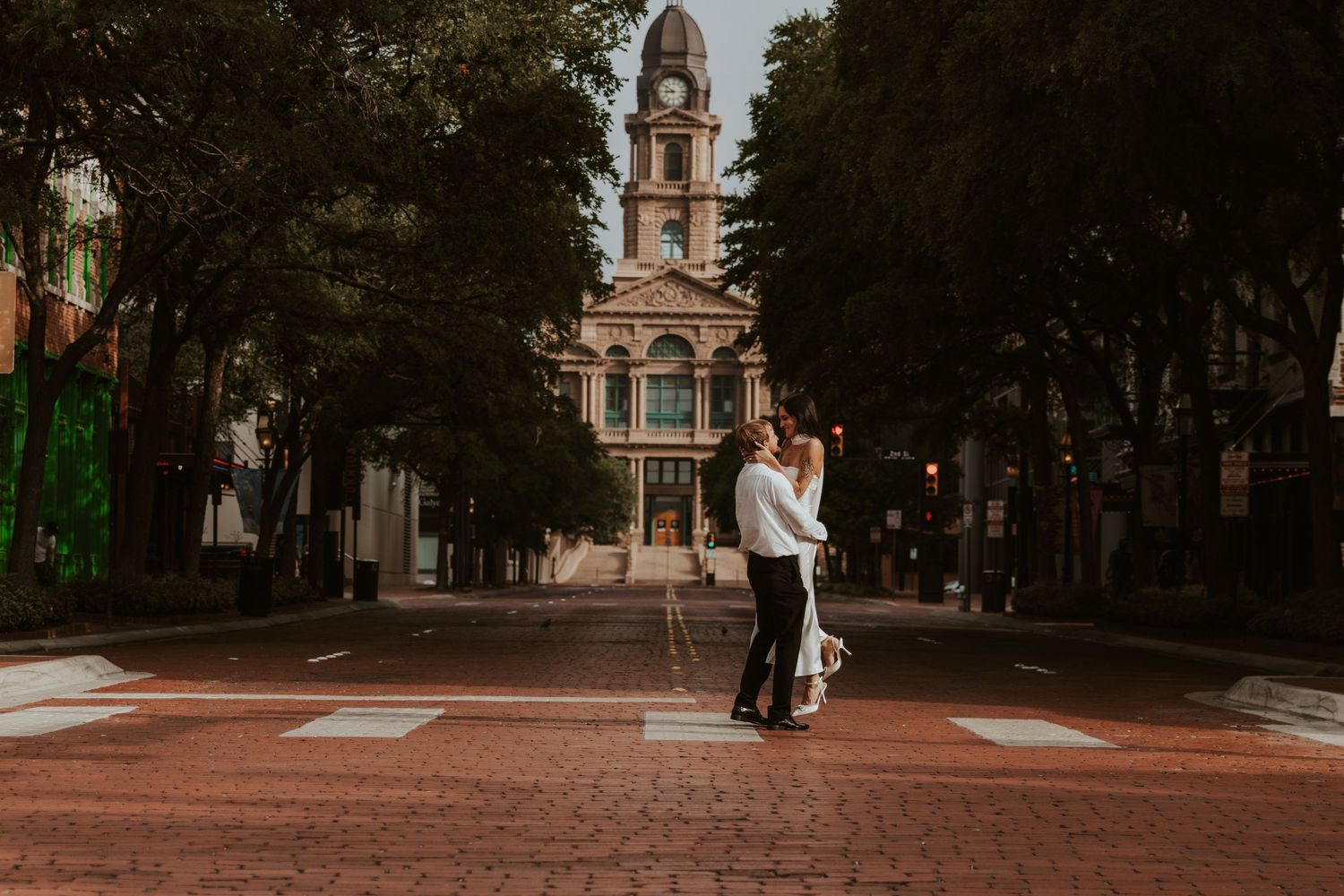 A person walks through a tree-lined campus pathway with historic brick buildings visible in the background. Engagement photographer DFW