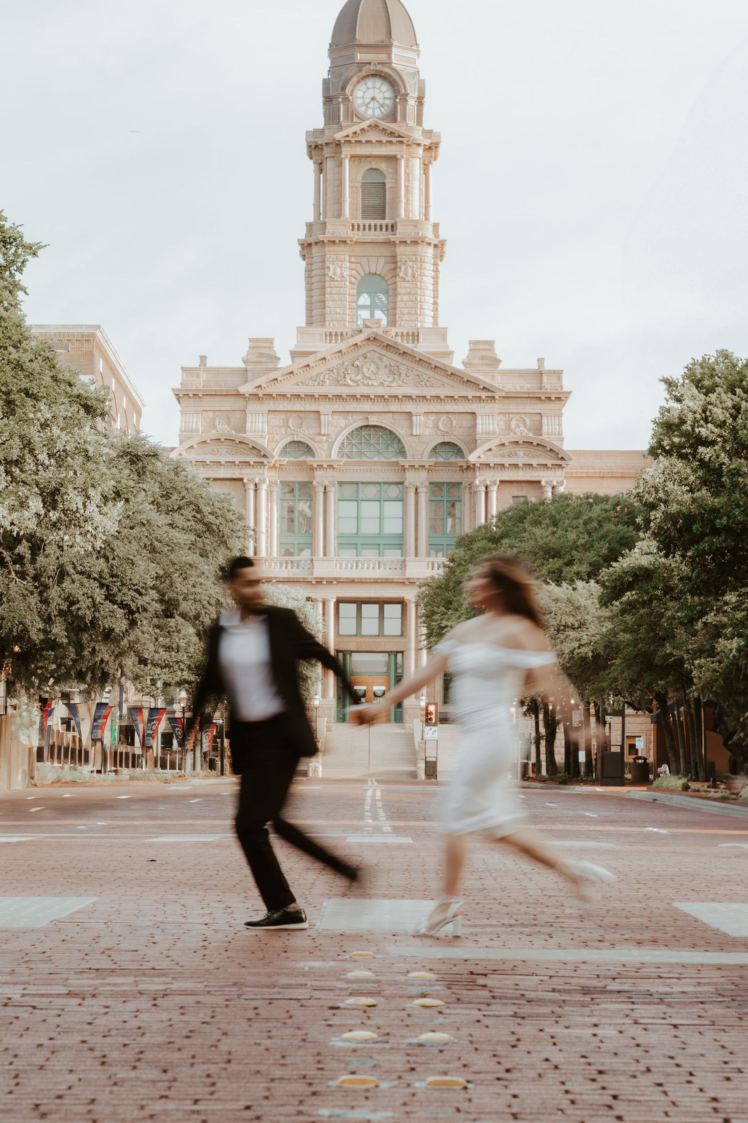 Motion blur captures two people walking in front of a historic brick clock tower building in soft sepia tones.