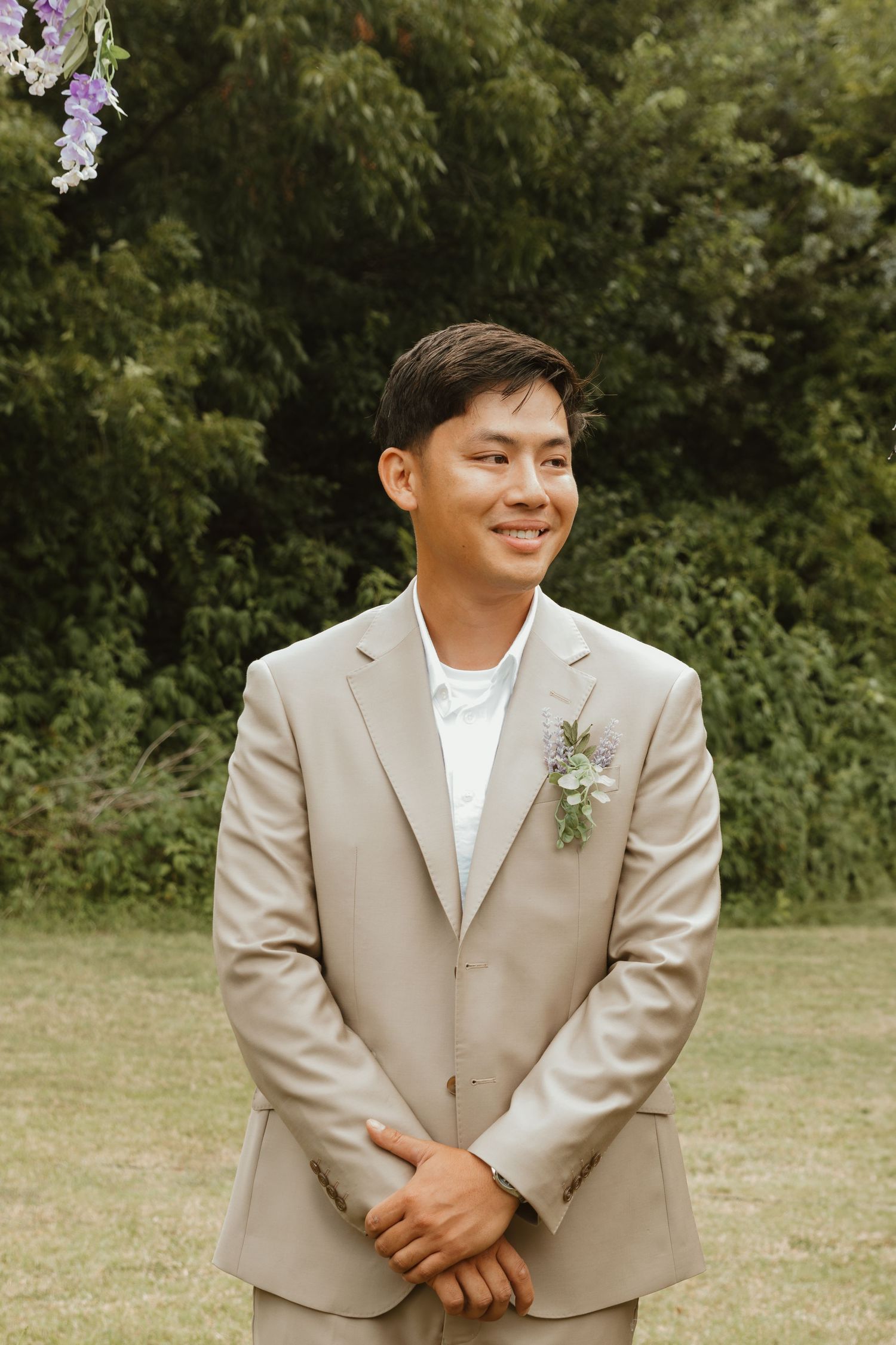 Groom in light beige suit with boutonniere smiles warmly while standing outdoors against lush green foliage.