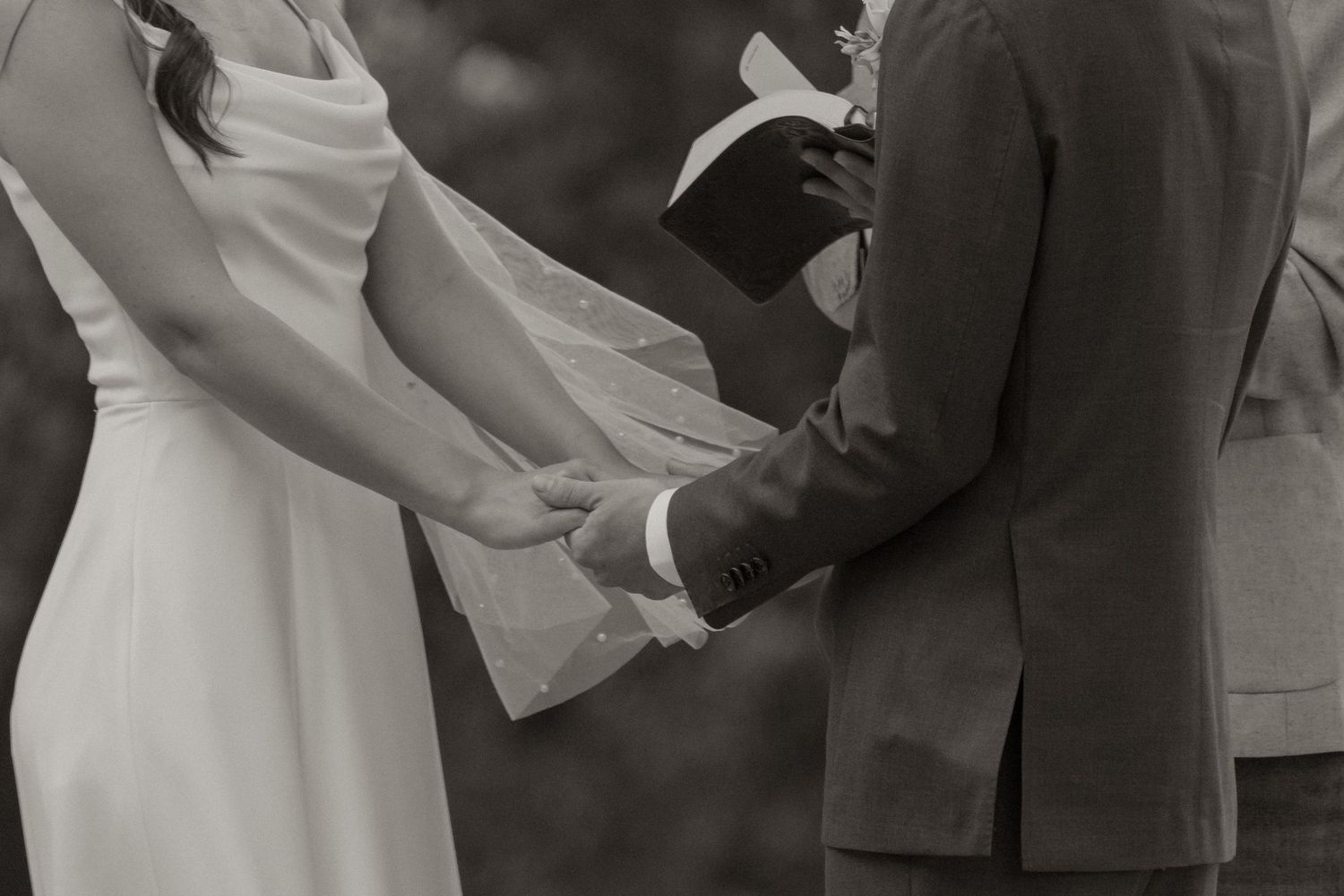 Black and white wedding photo of bride and groom holding hands during outdoor ceremony with officiant.