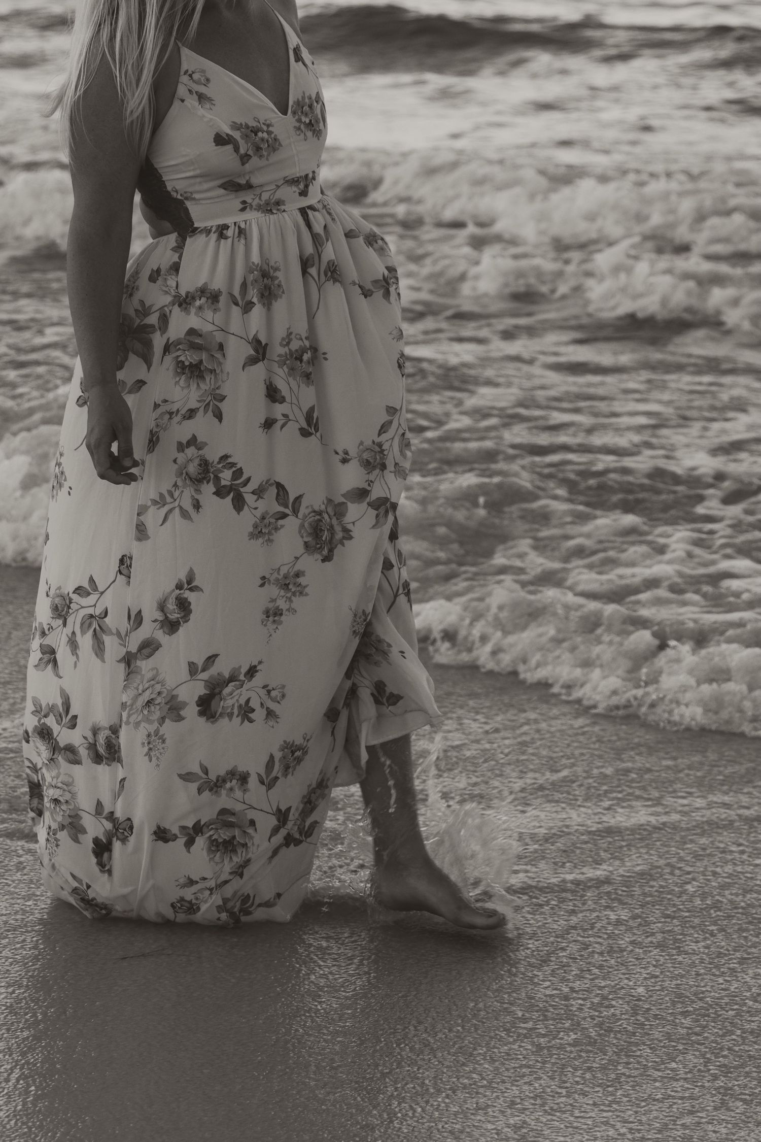 Woman in floral maxi dress standing barefoot on sandy beach with ocean waves, black and white photo.