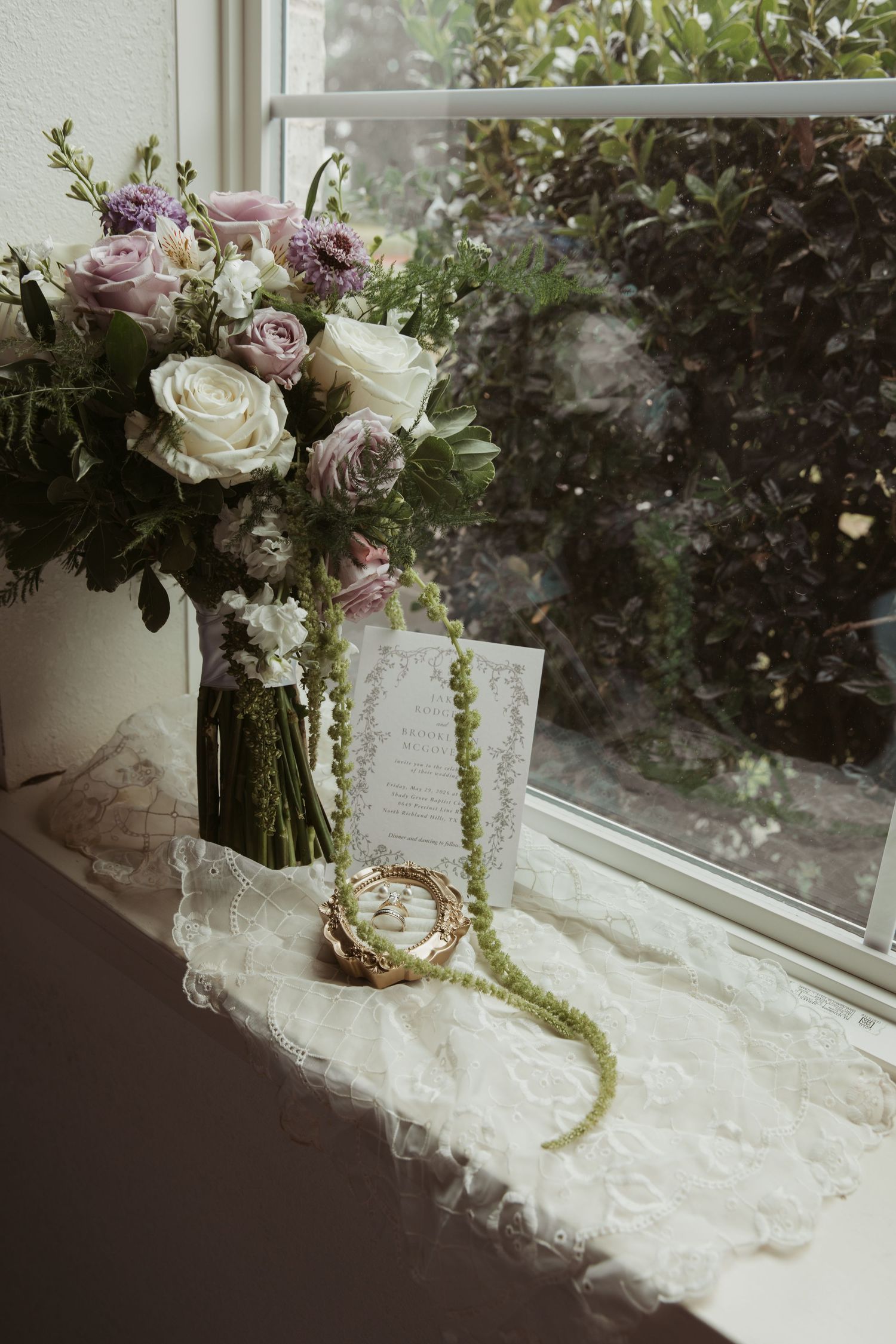 Elegant bridal bouquet of white and mauve roses on lace with wedding rings and greenery by a window.