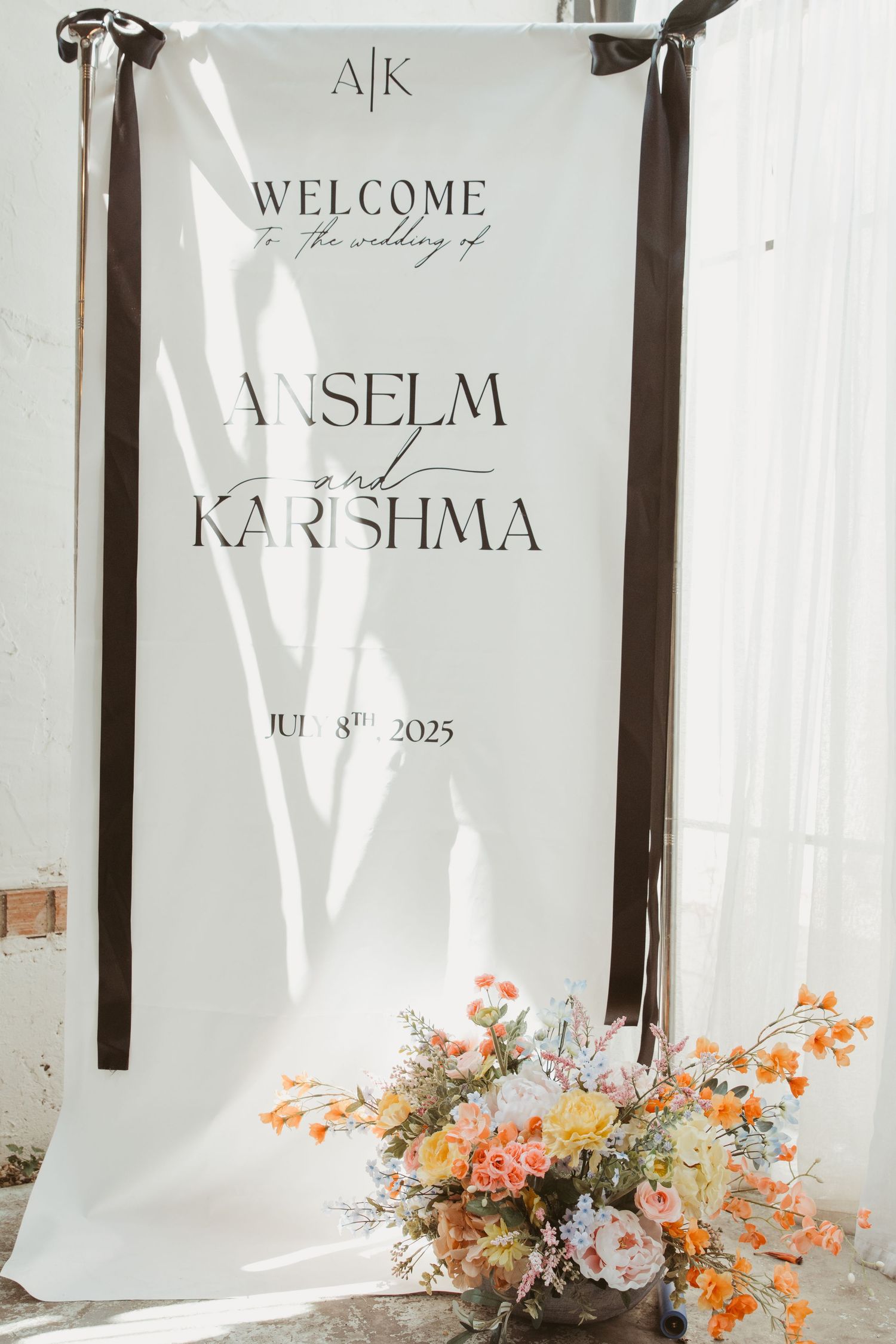 Elegant wedding welcome sign with white backdrop and orange floral arrangement featuring names Anselm and Karishma.