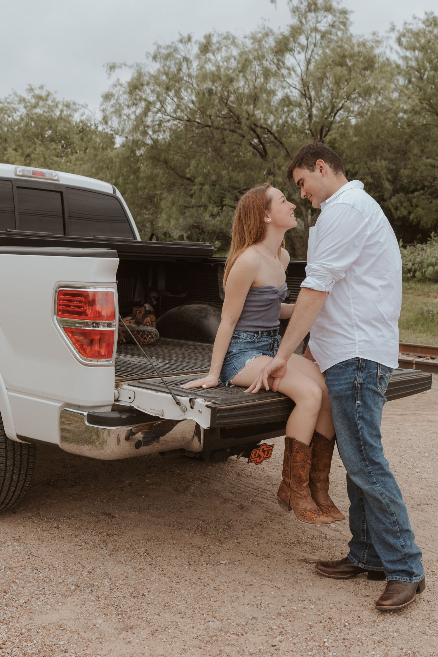 Two young people share a romantic moment while sitting on the tailgate of a white pickup truck at dusk.