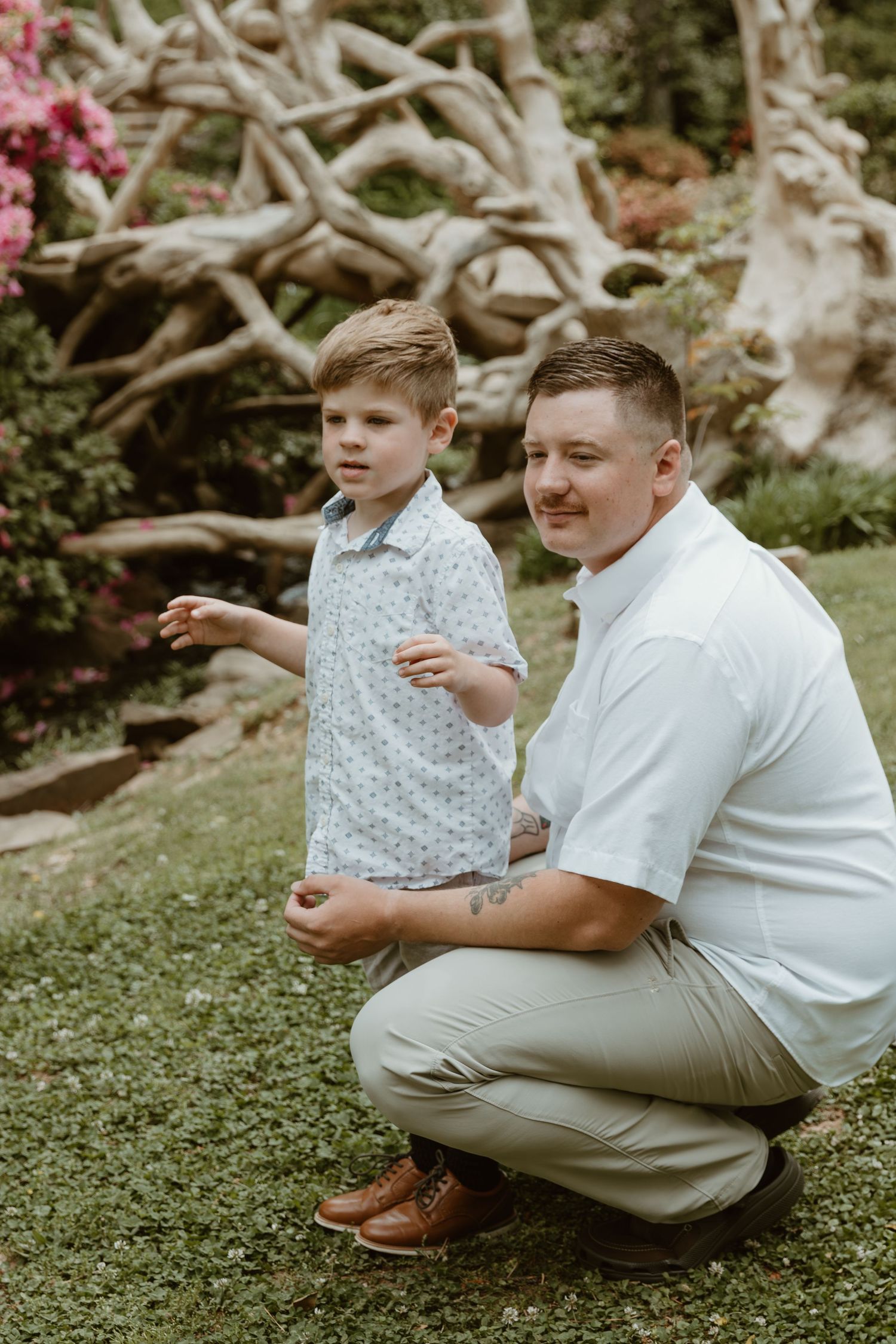 A tender outdoor family moment captured in a garden with sculptural driftwood and pink flowers in the background.