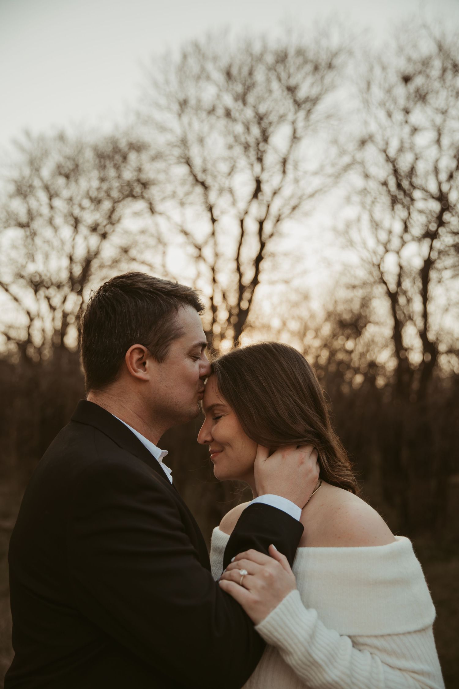 Romantic couple embracing at sunset among bare winter trees, woman wearing engagement ring in white sweater.