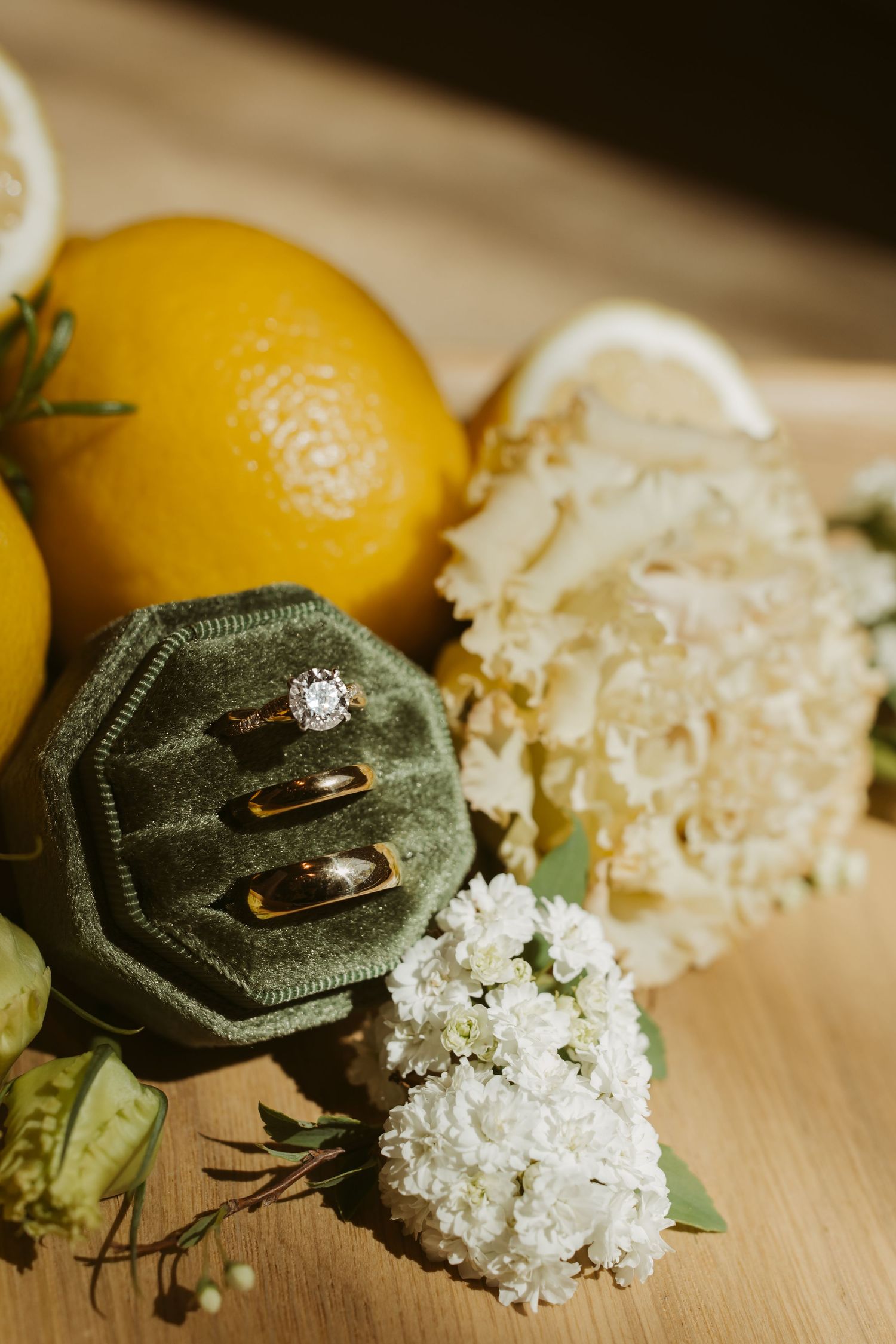 Wedding rings in green velvet box with diamond engagement ring, surrounded by lemons and white flowers.