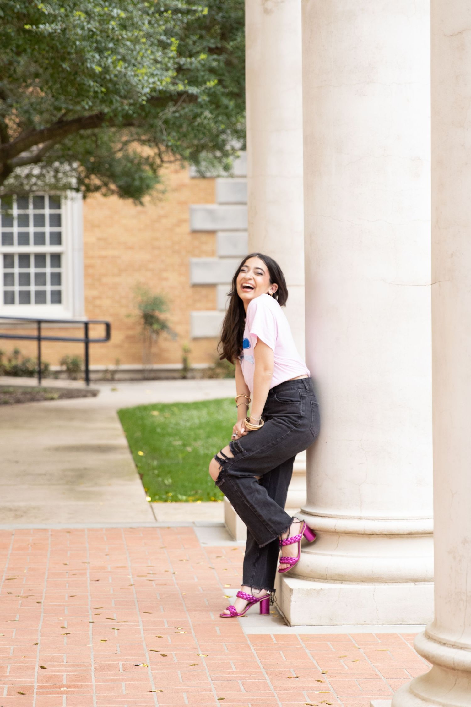 Playful pose leaning against a white column in pink heels and dark pants on a university campus.