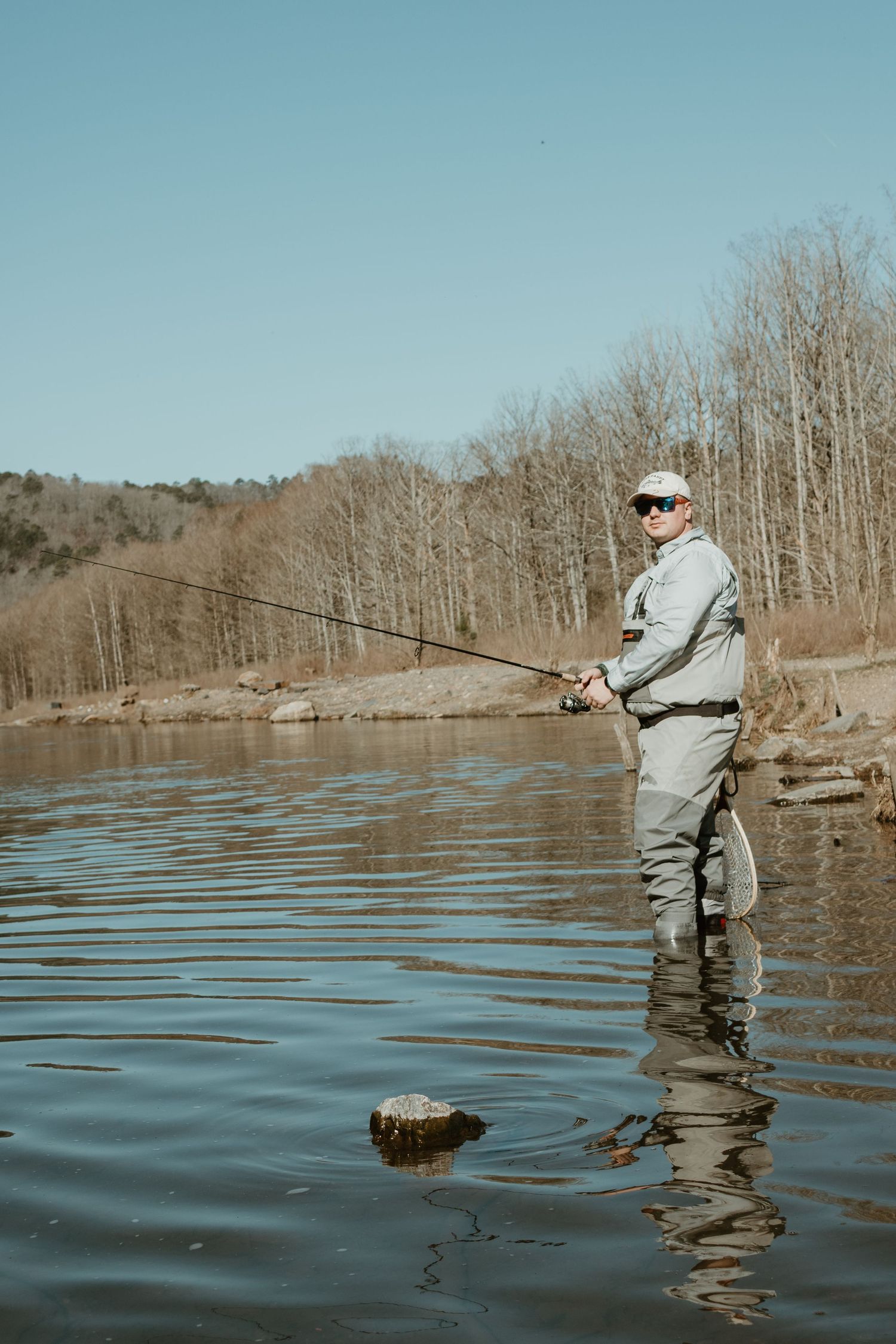 An angler in waders casts a fishing line into a calm lake surrounded by bare winter trees.