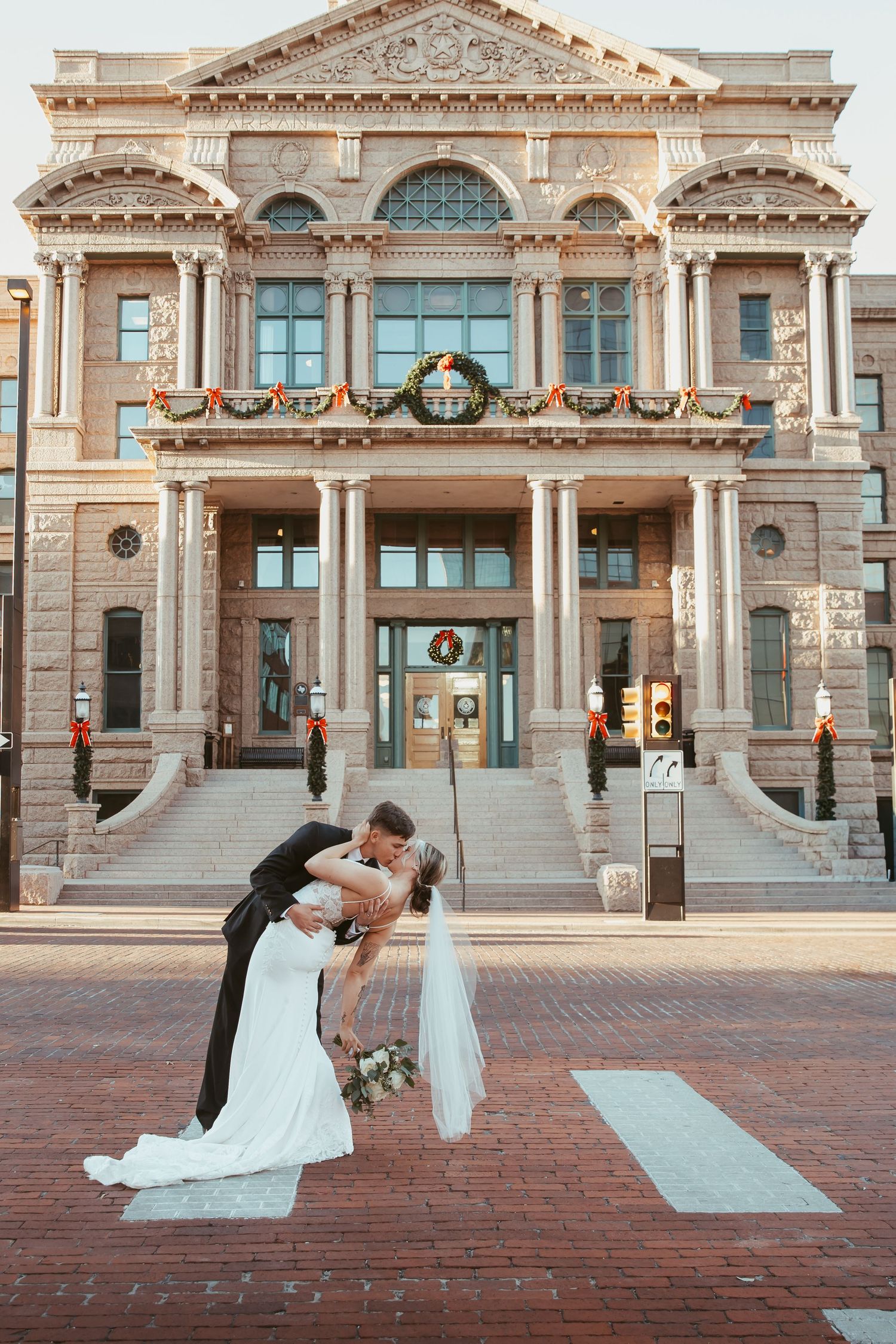 Two brides in white wedding dresses share a romantic moment on brick steps of a historic courthouse building.