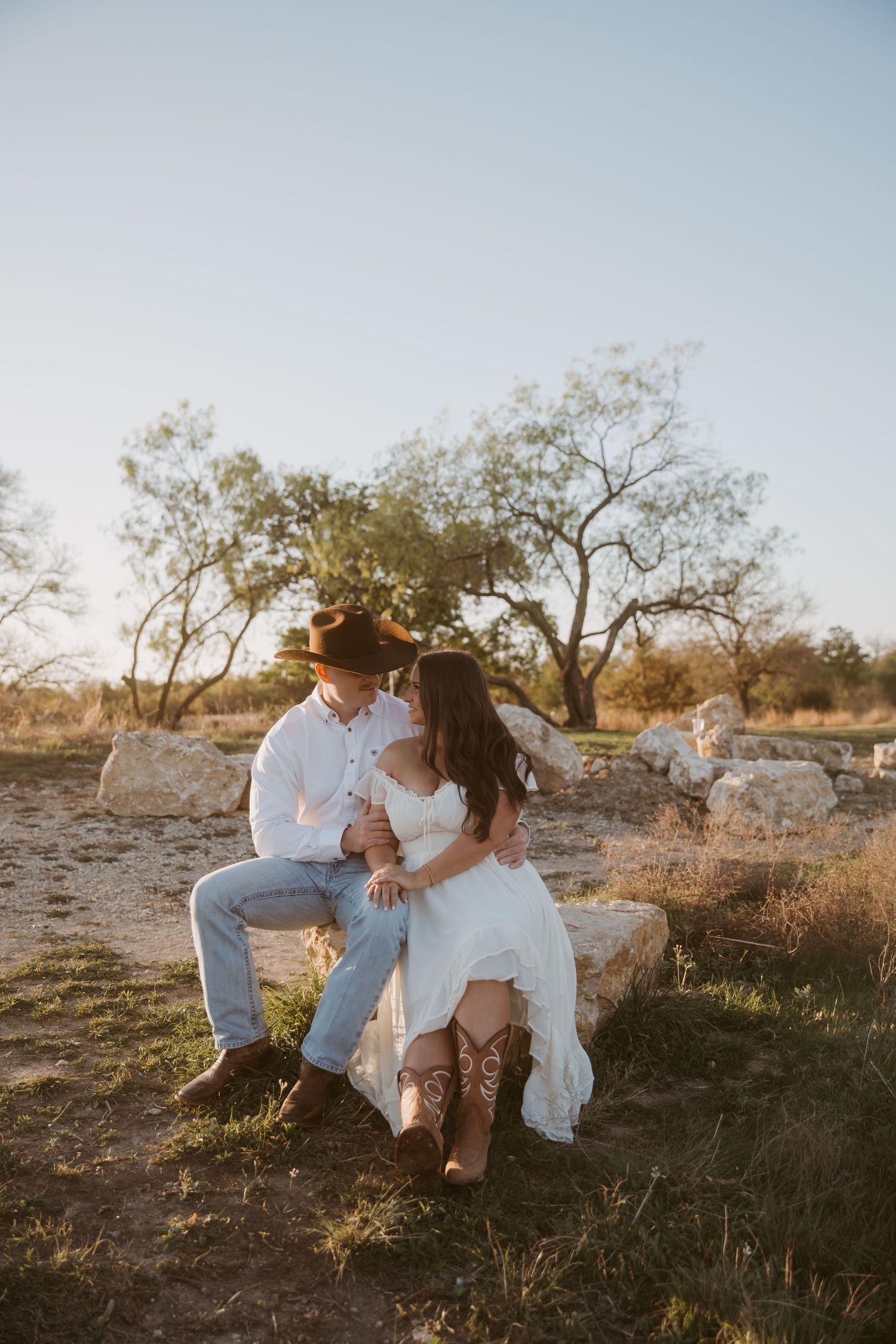 Couple sitting on stone in rustic outdoor setting, man in cowboy hat and woman in white dress with cowboy boots.
