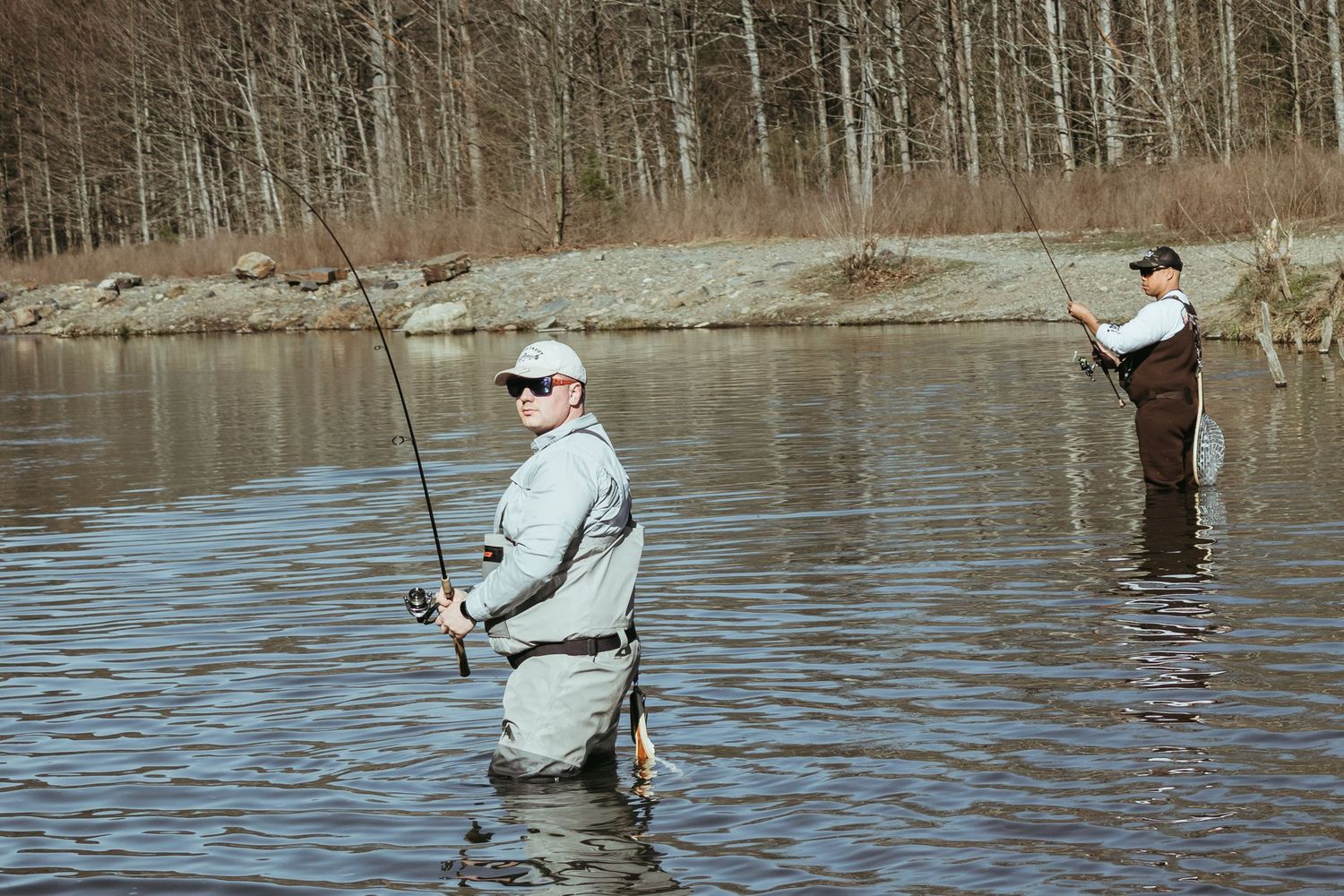Two anglers in waders fish together in a tranquil lake surrounded by trees.