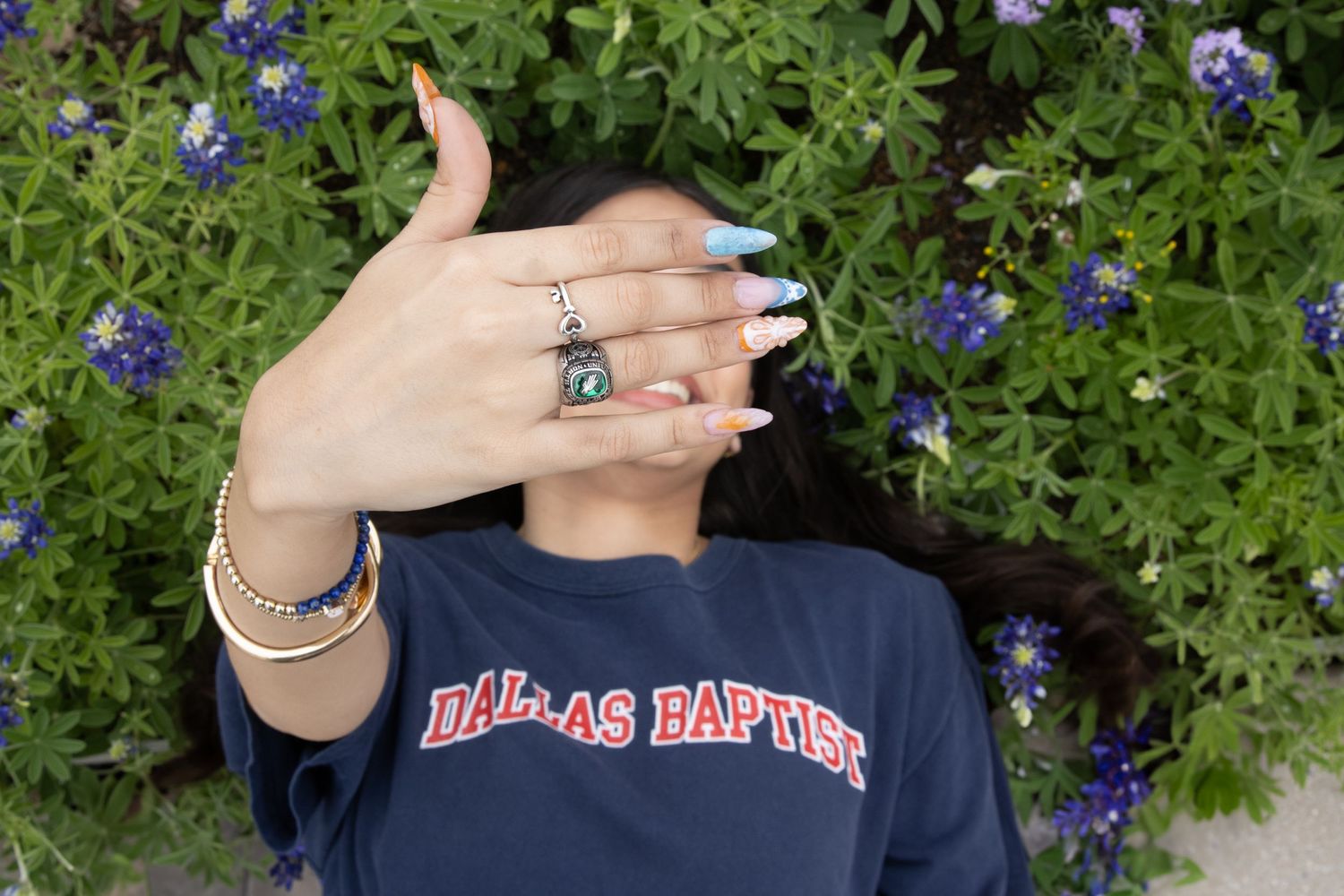 A hand with silver rings and blue nail polish held up against bluebonnet flowers and a Dallas Baptist University shirt.