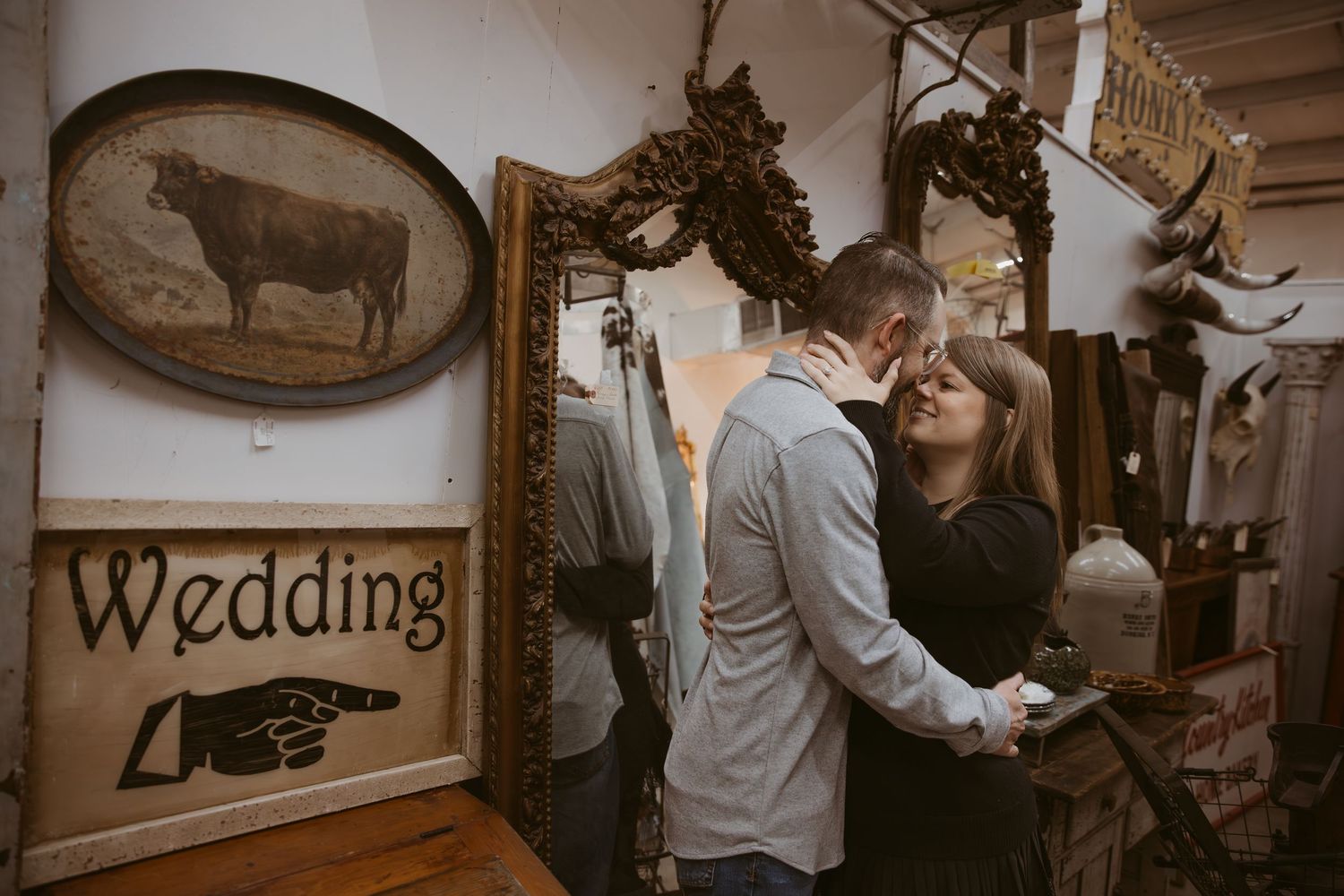 A romantic couple embraces in a rustic antique shop with vintage decor and a wedding sign on the wall.
