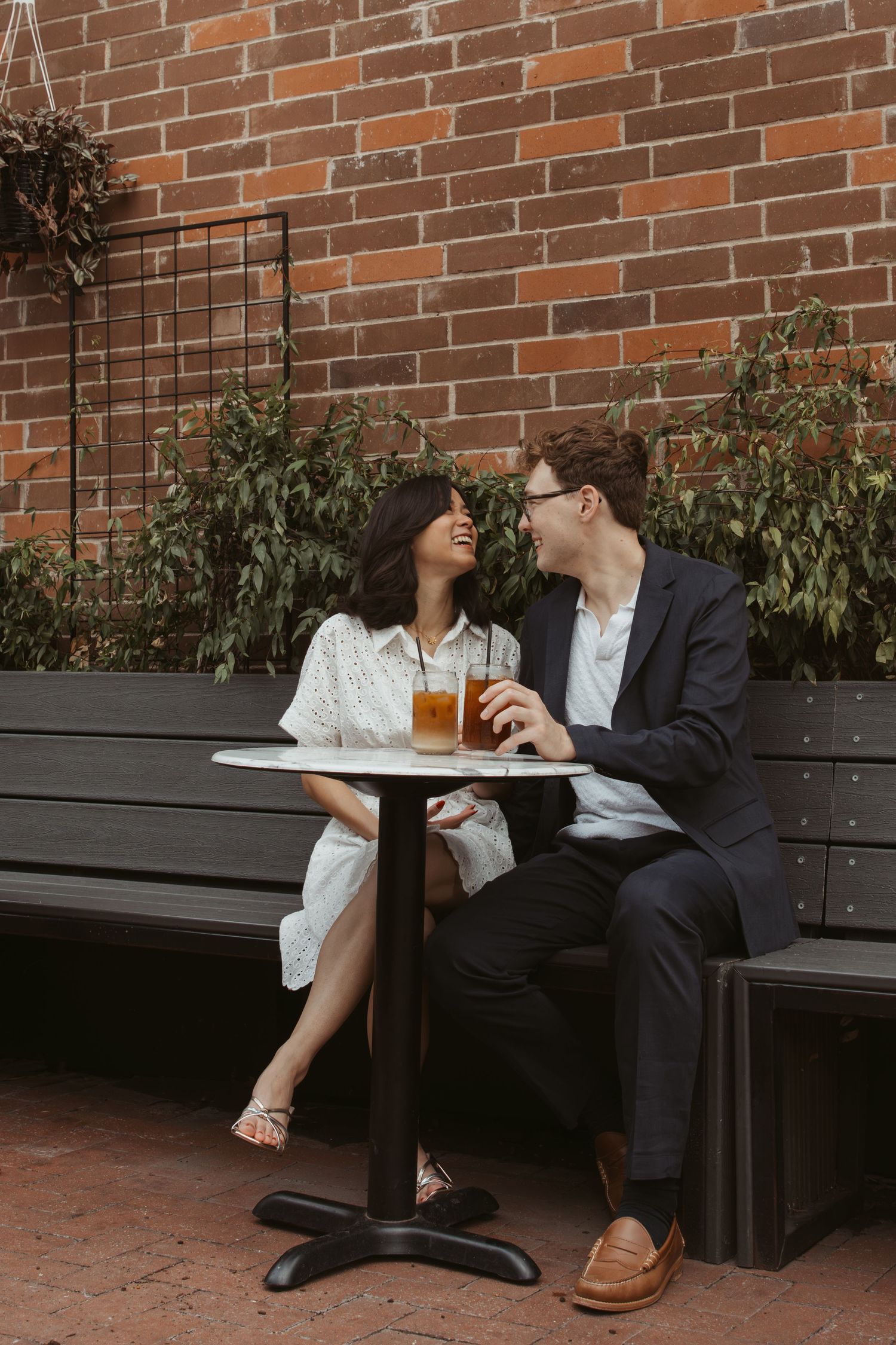 A couple shares a romantic moment on a bench against a brick wall with greenery.