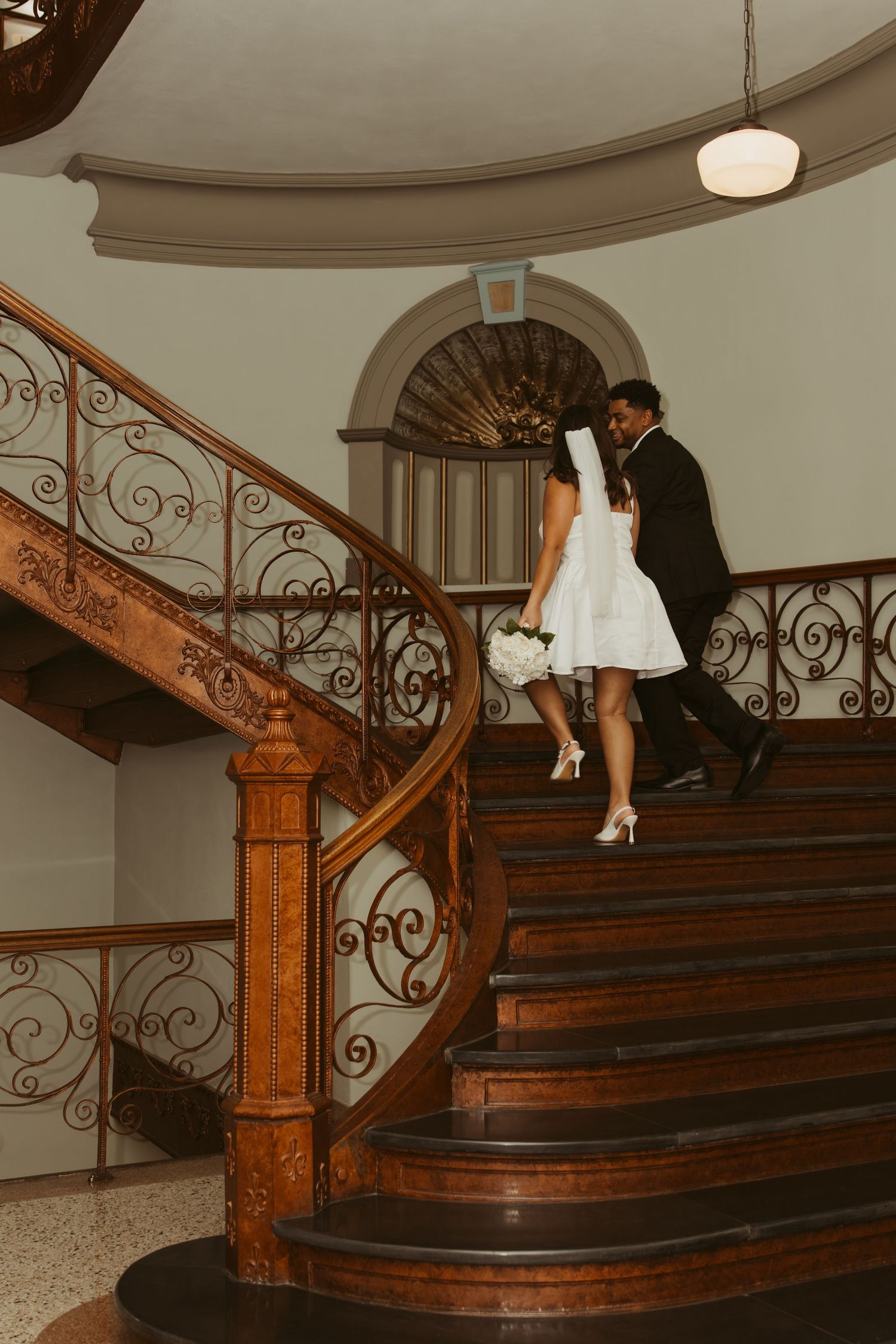 A couple in formal wear poses on a grand wooden staircase with ornate iron railings. Wedding Photographer DFW