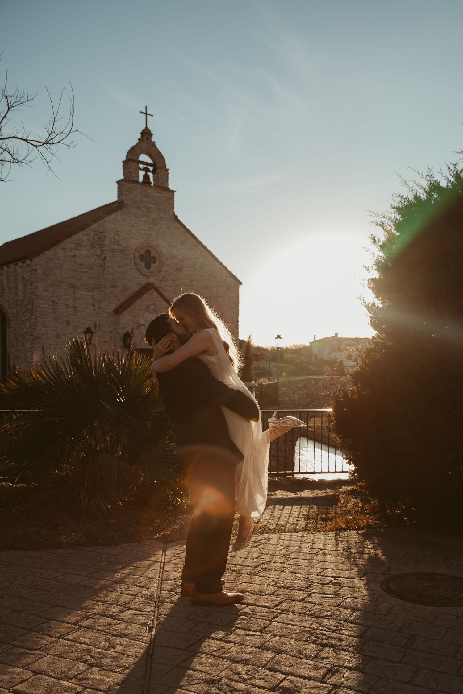 A silhouetted couple dances in front of a rustic church at sunset, casting long shadows on the cobblestone ground.