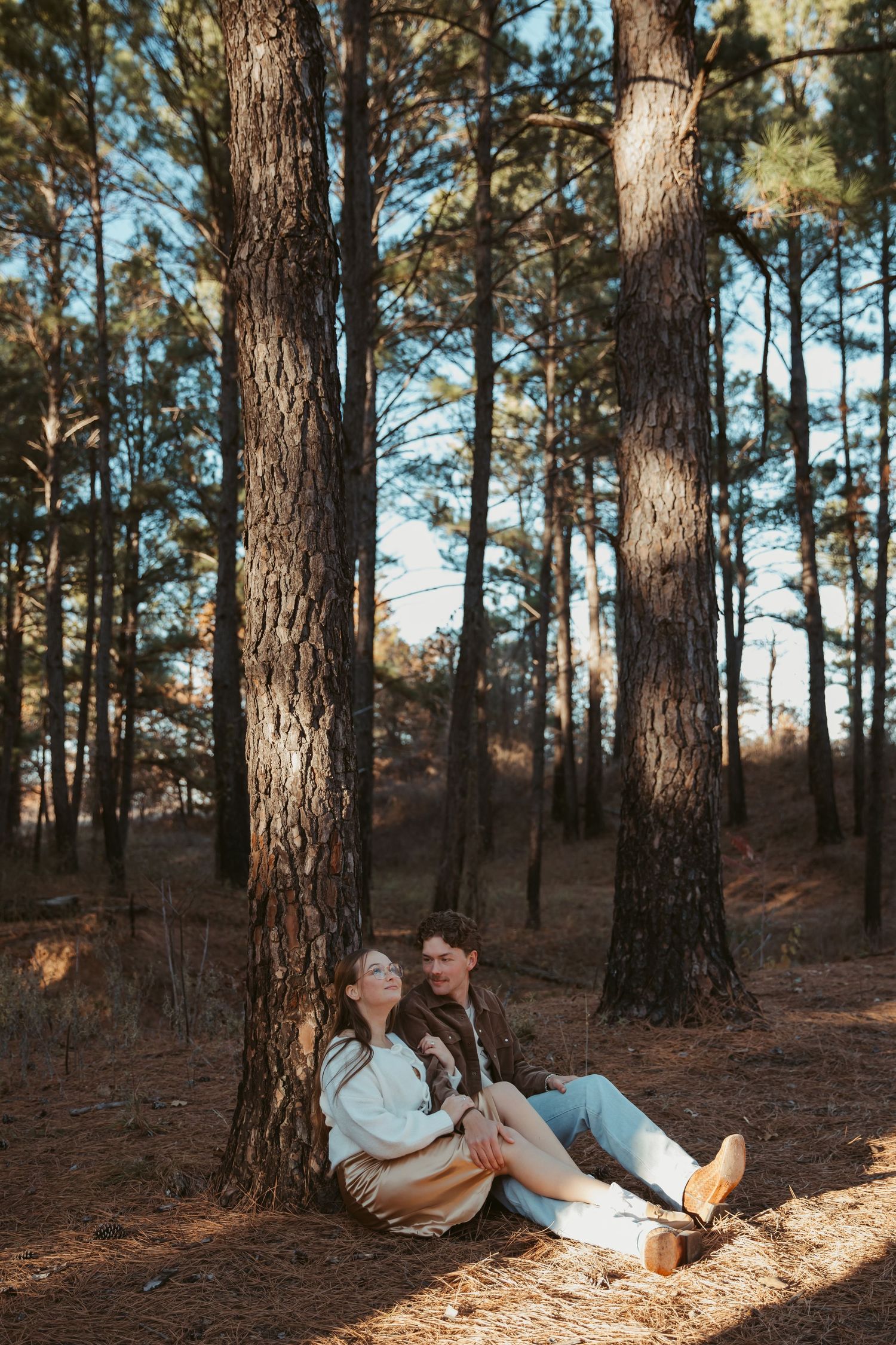 Couple relaxing on pine needle covered forest floor surrounded by tall trees in warm afternoon light. engagement photos Fort Worth