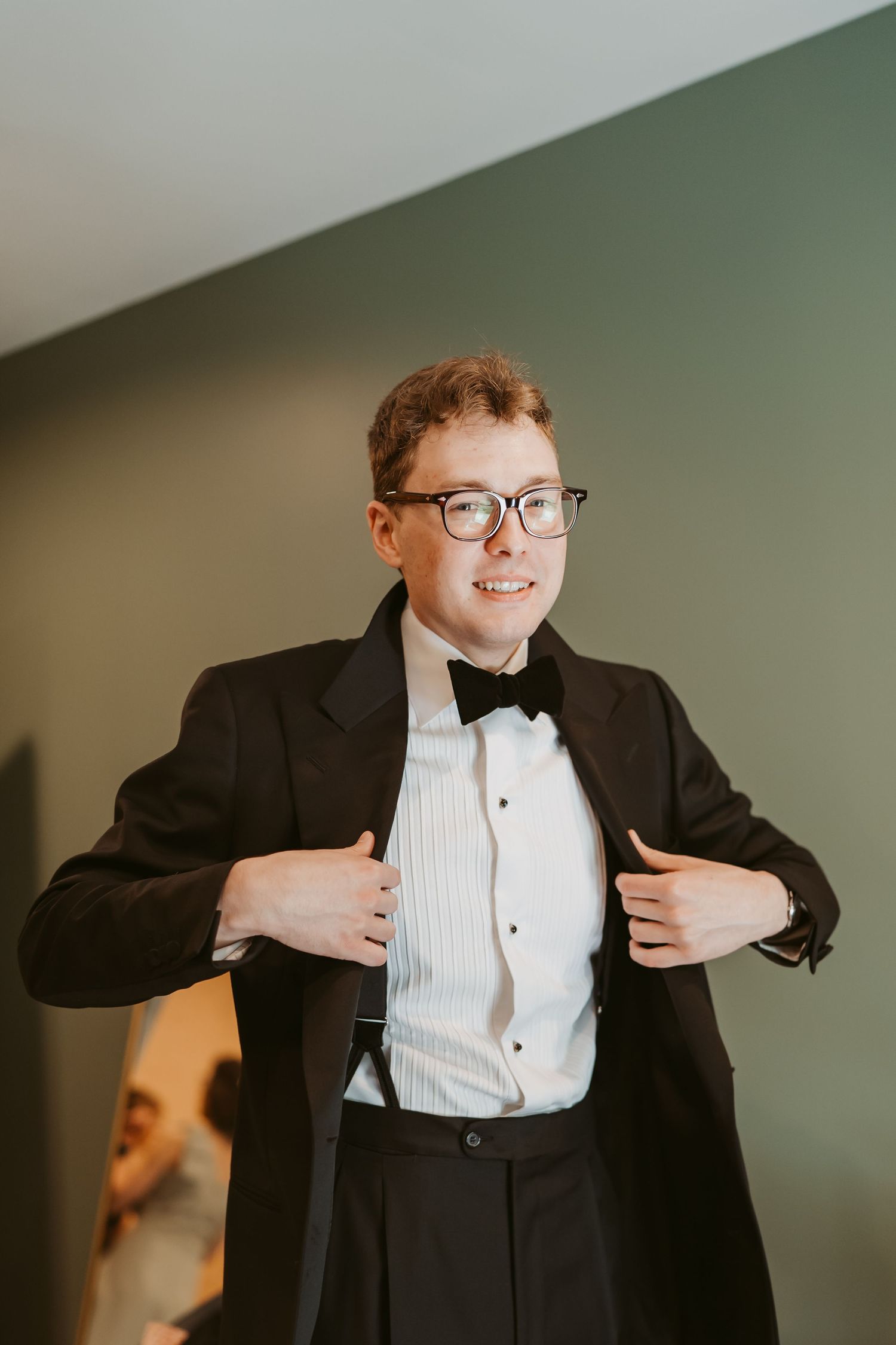 A young man in a black tuxedo and bow tie adjusts his jacket lapels while smiling, wearing glasses.