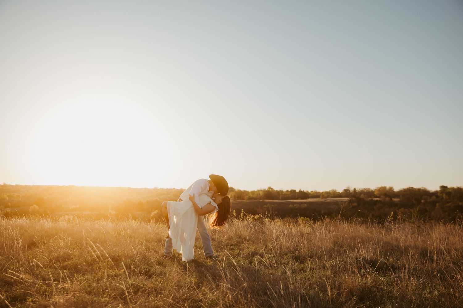 Couple sharing a romantic dip kiss in a golden sunlit field during sunset, surrounded by tall grass.