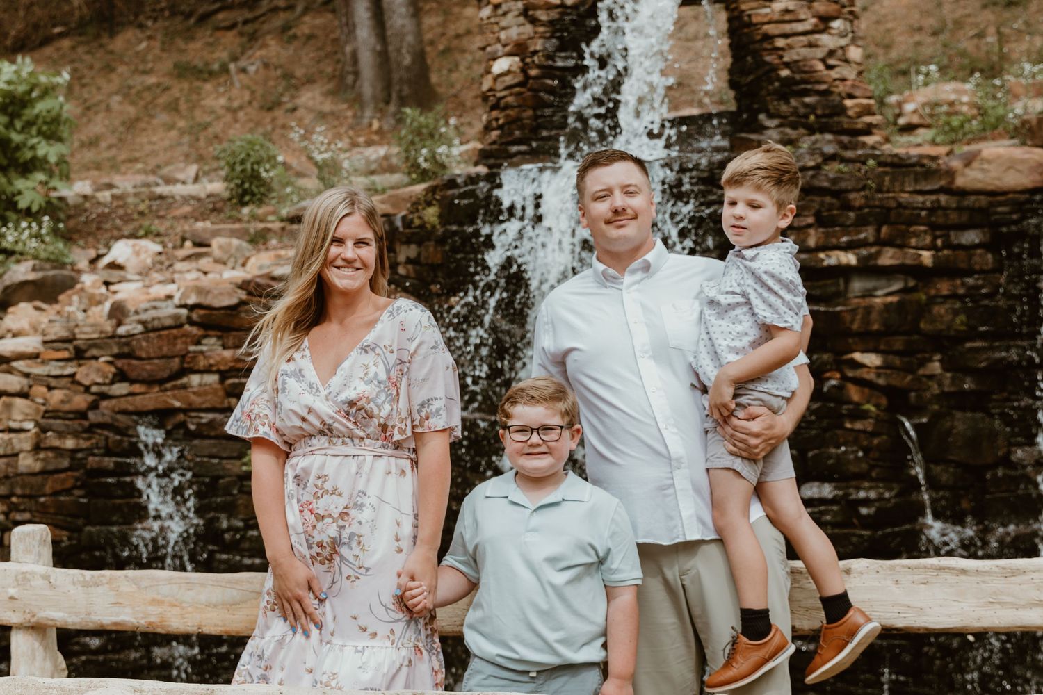 A family poses together in front of a cascading waterfall on a summer day while standing at a wooden railing.