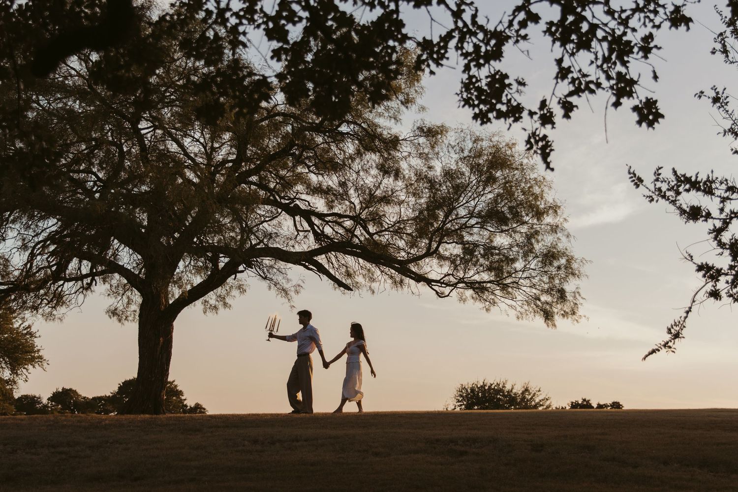 Silhouetted figures hold hands against a sunset sky with tree branches framing the scene.