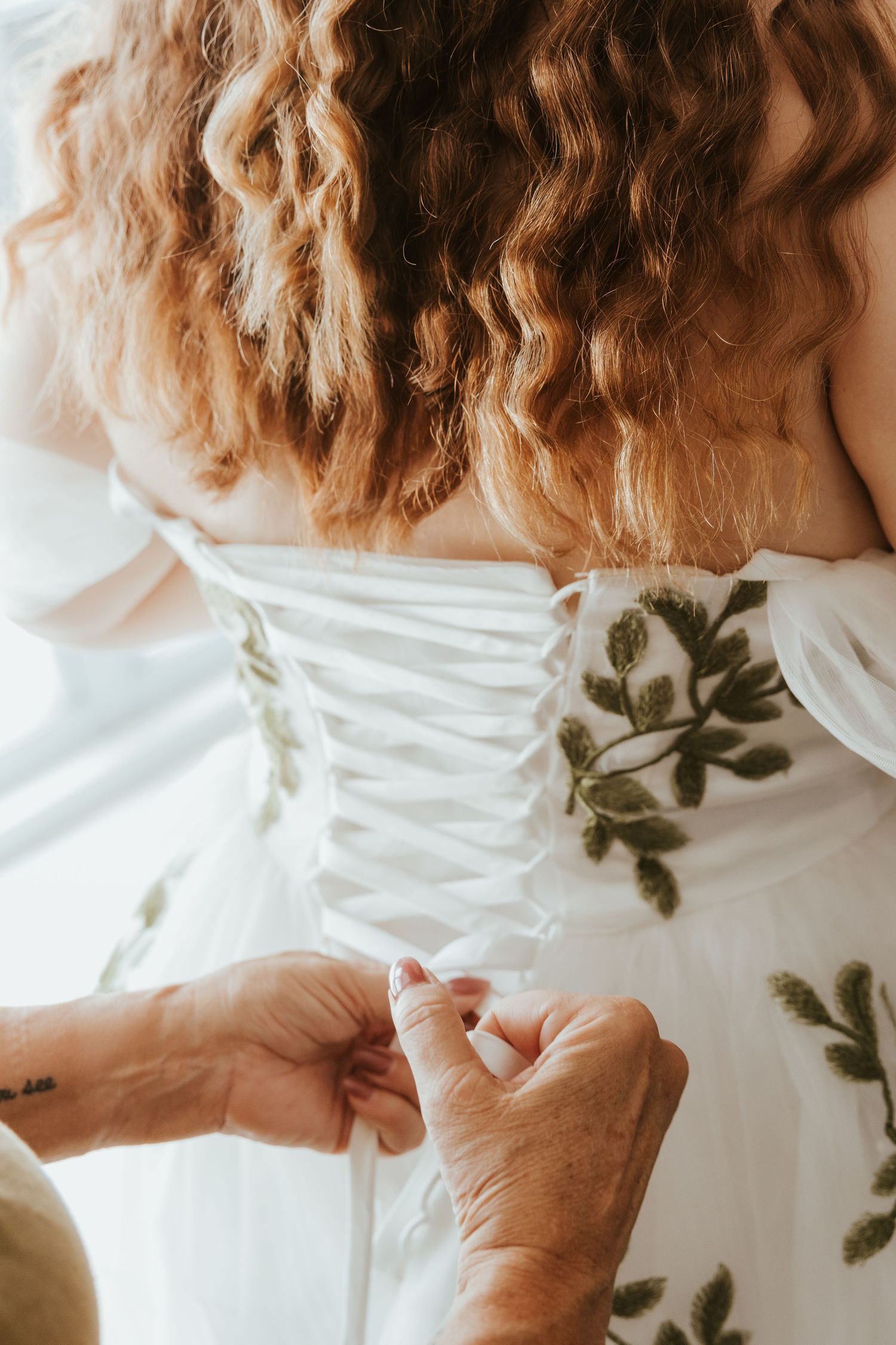 Close-up view of hands fastening a white wedding dress corset with green foliage accent.