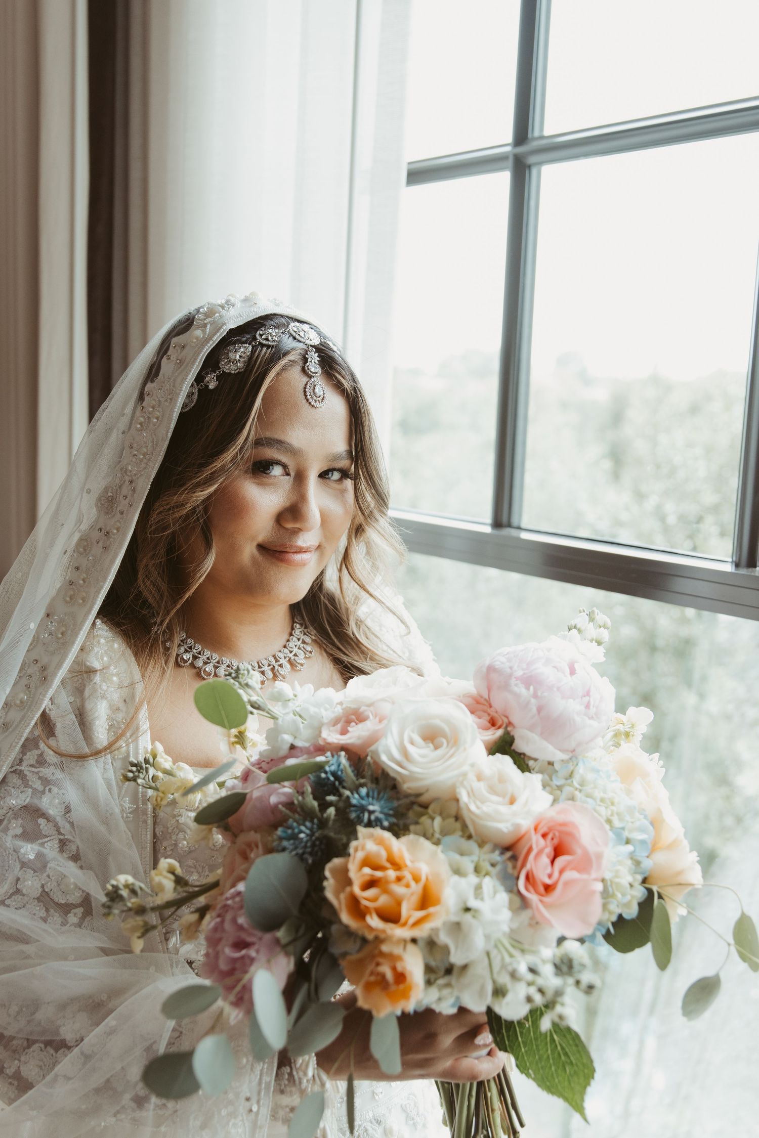 Elegant bridal portrait featuring a lace dress and peach-toned floral bouquet by a bright window.