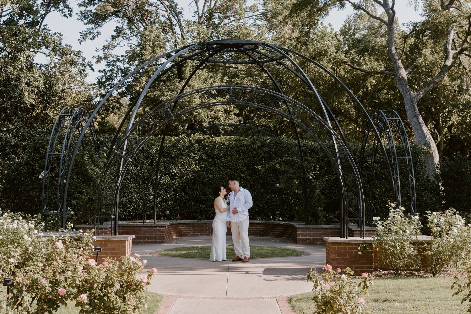 A couple embraces under an arched metal trellis in a garden setting surrounded by trees and flowering plants.