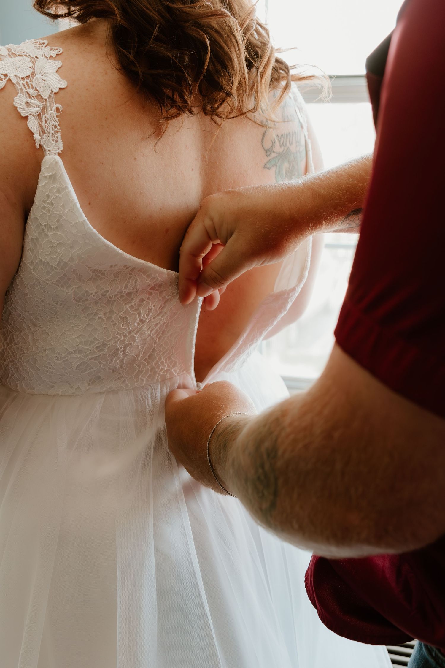 Someone helps zip up a delicate white lace wedding dress with intricate details.