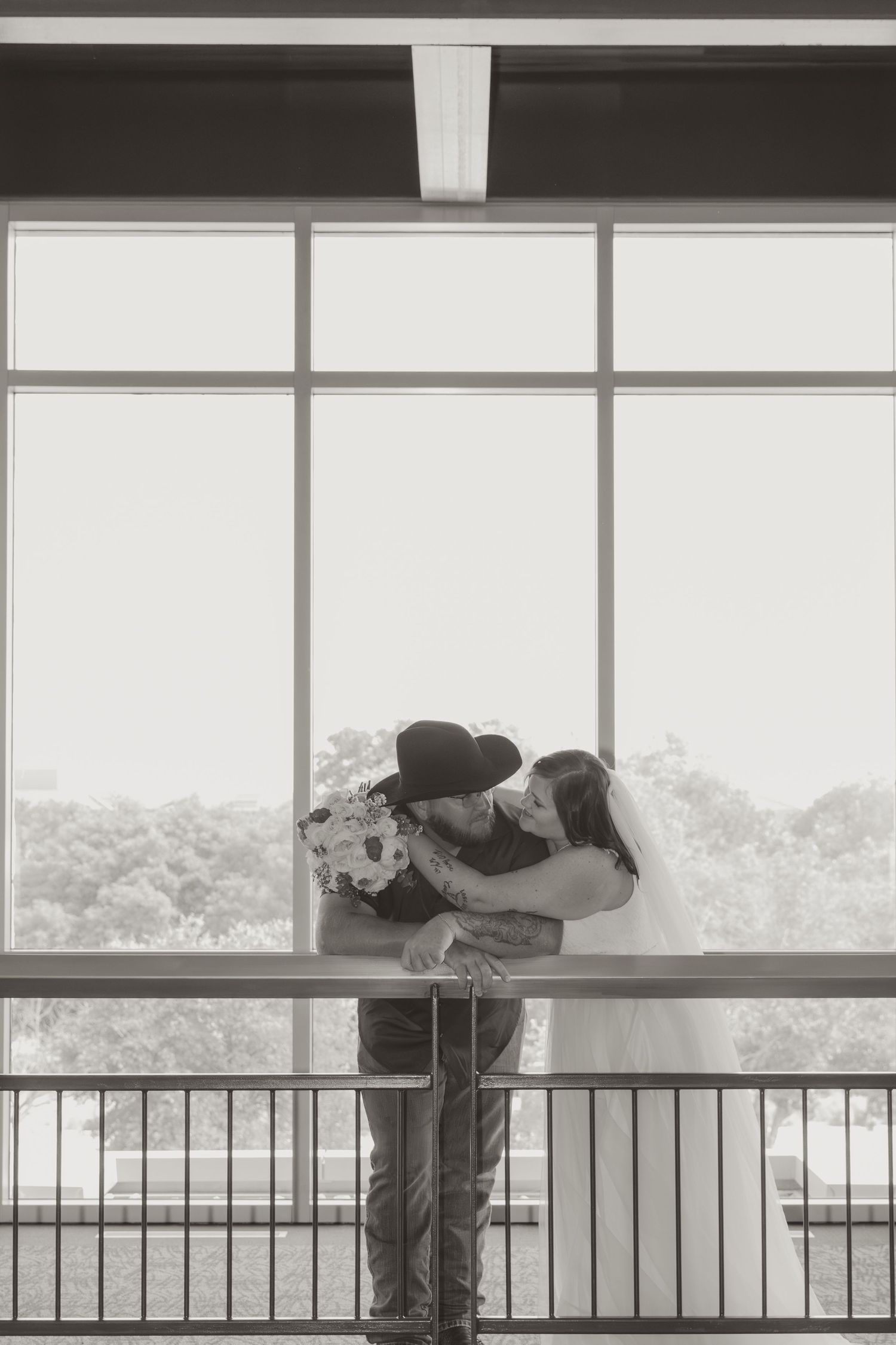 A romantic black and white silhouette of a couple embracing by a window railing with trees visible in background.
