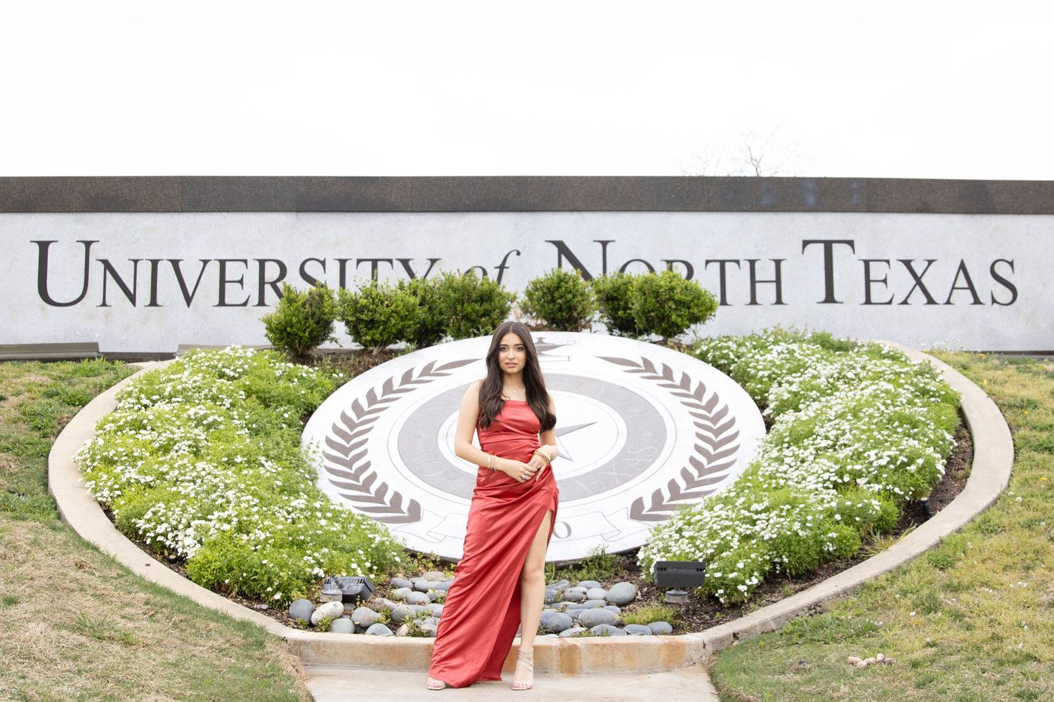Graduate in red gown posing at the University of North Texas sign with white flowers.