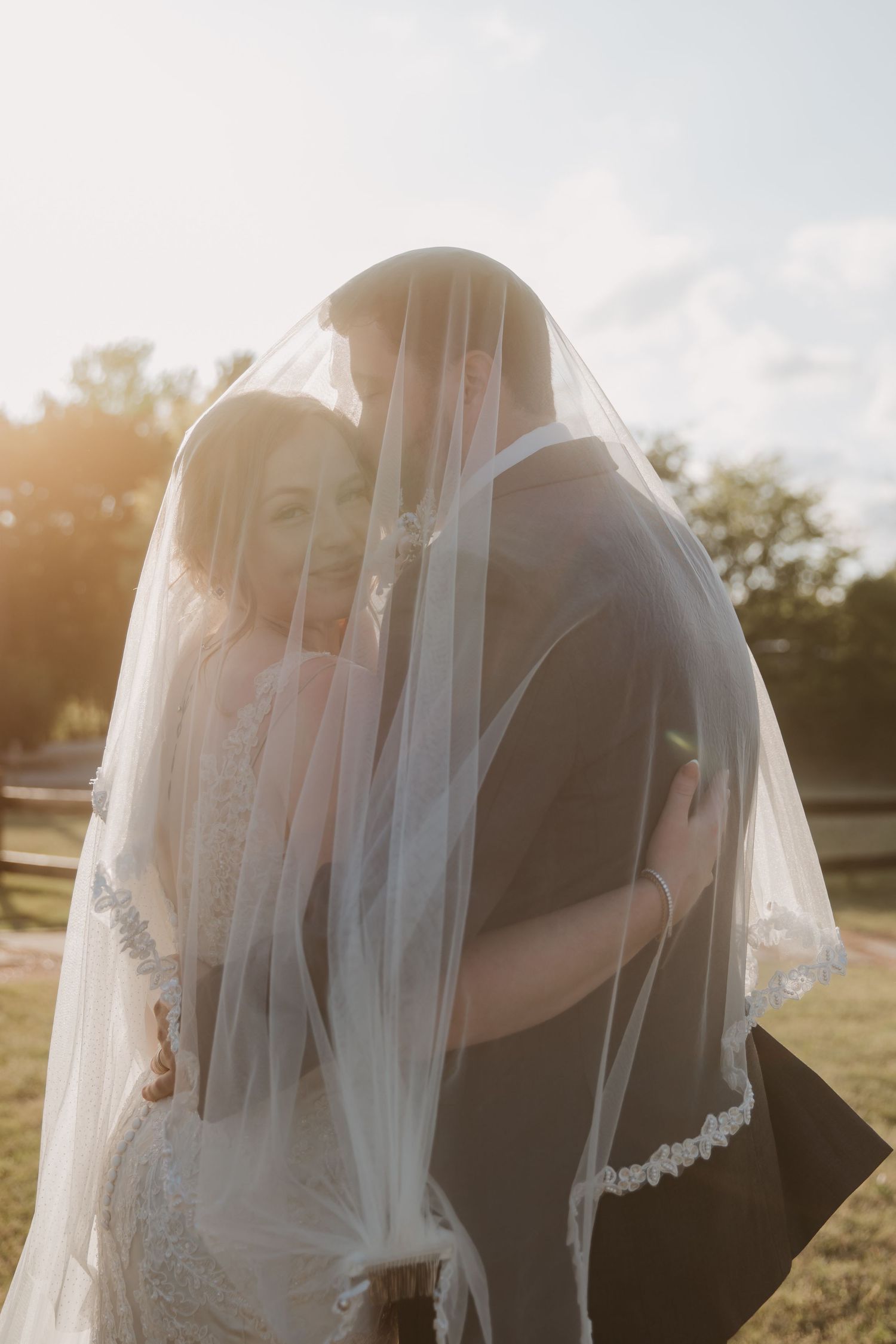 Bride and groom sharing a romantic kiss under a flowing white wedding veil in golden sunset light outdoors.