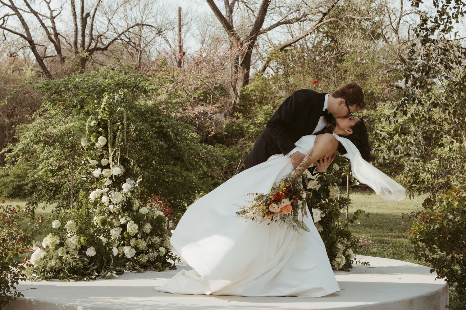 A groom dips his bride for a romantic kiss outdoors, her white gown and floral bouquet flowing gracefully.