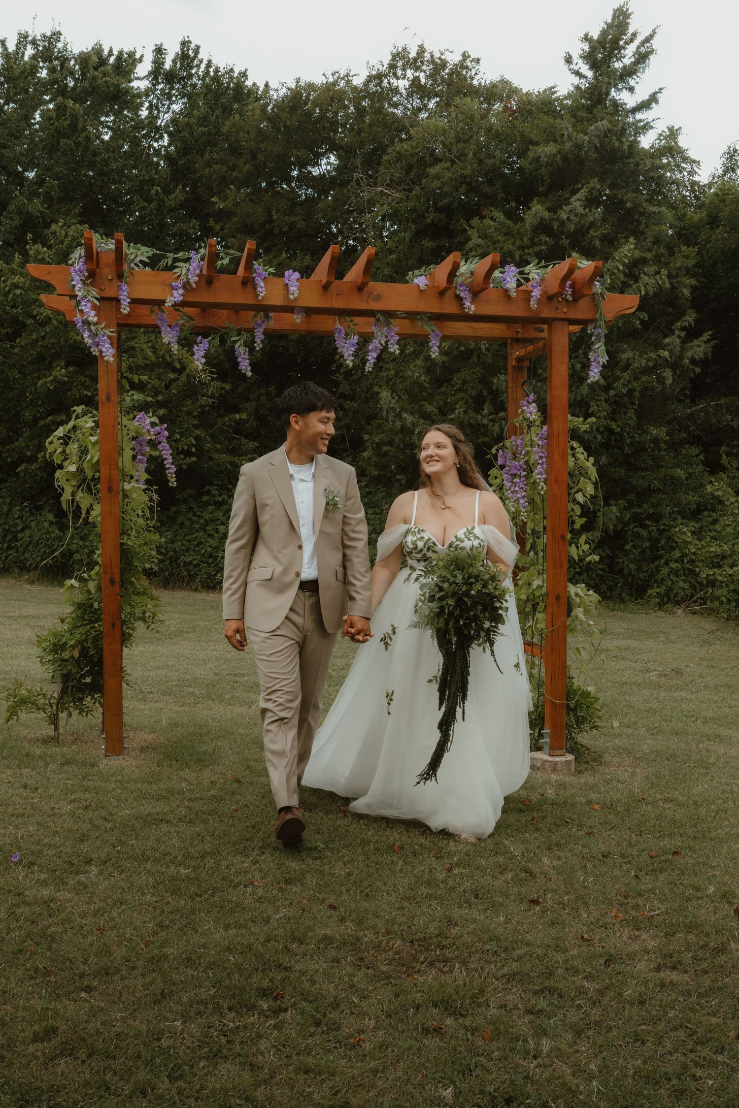 A newlywed couple walks together through a grassy field under wooden pergolas with floral decorations.