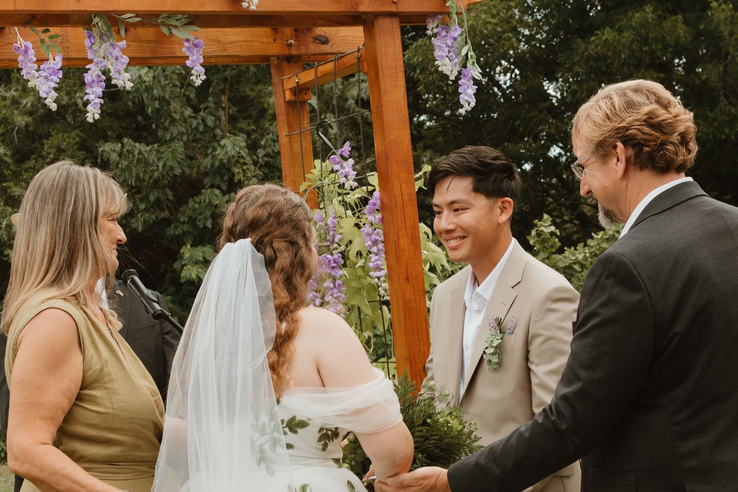 Outdoor wedding ceremony under a wooden arch decorated with purple flowers.