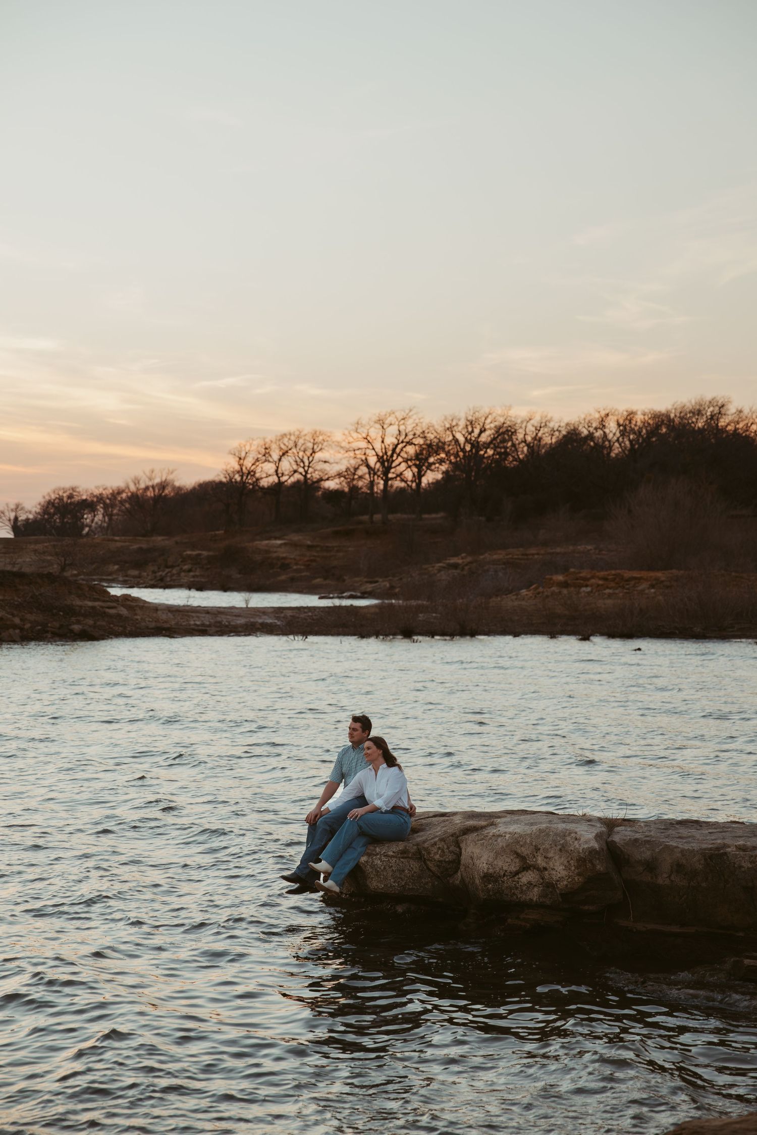 Couple sitting together on rocks by a peaceful lake at sunset, with bare trees lining the horizon.
