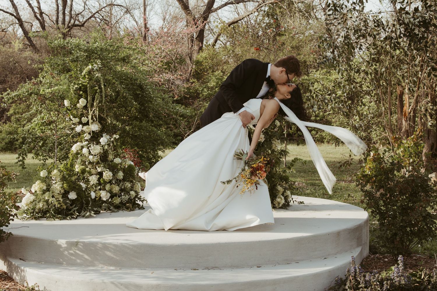 Groom dips bride in white wedding dress for romantic kiss surrounded by lush garden florals.