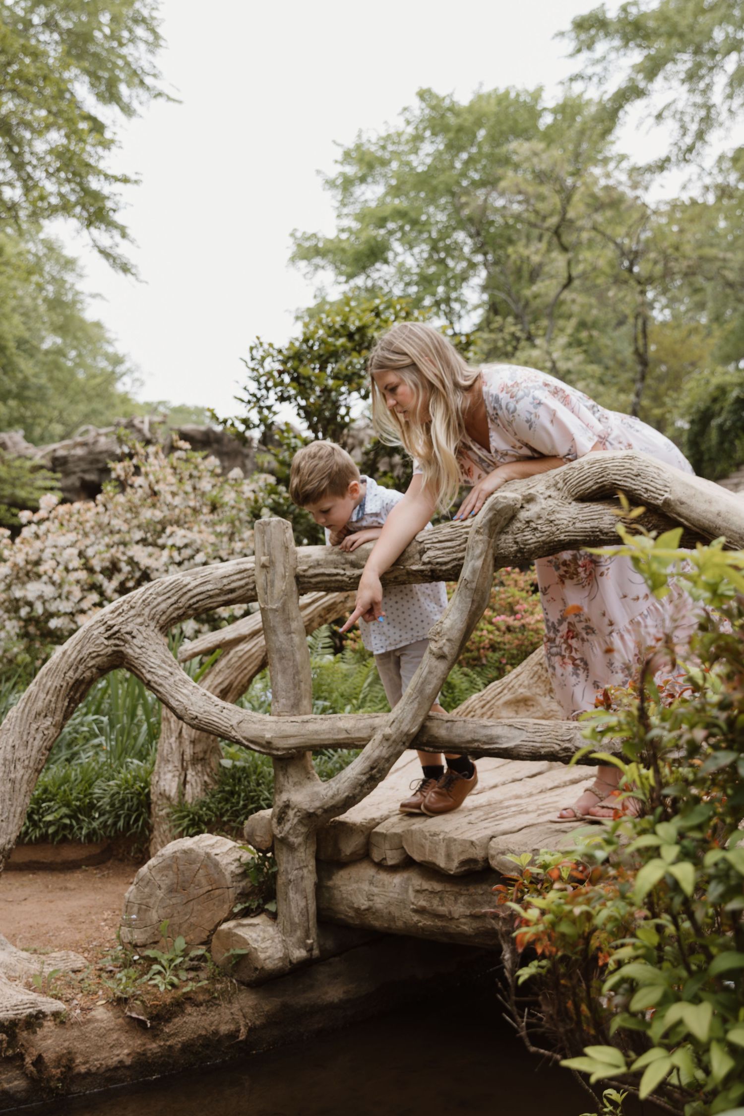 A person in a floral dress lies across a rustic wooden bridge in a garden setting during a nature photoshoot.