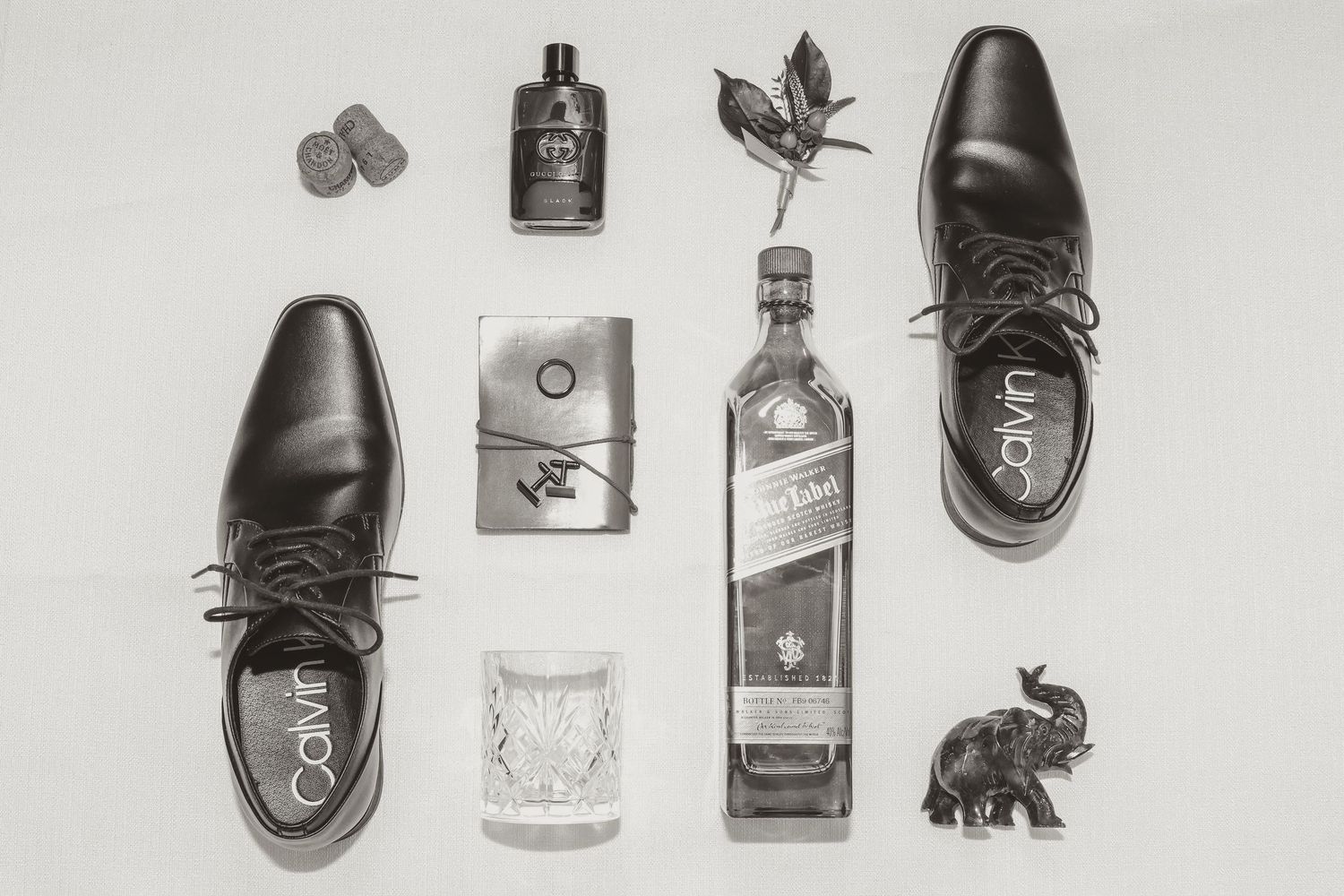 A black and white flat lay arrangement of dress shoes, whiskey bottle, perfume, wallet, drinking glass and decorative elephant.