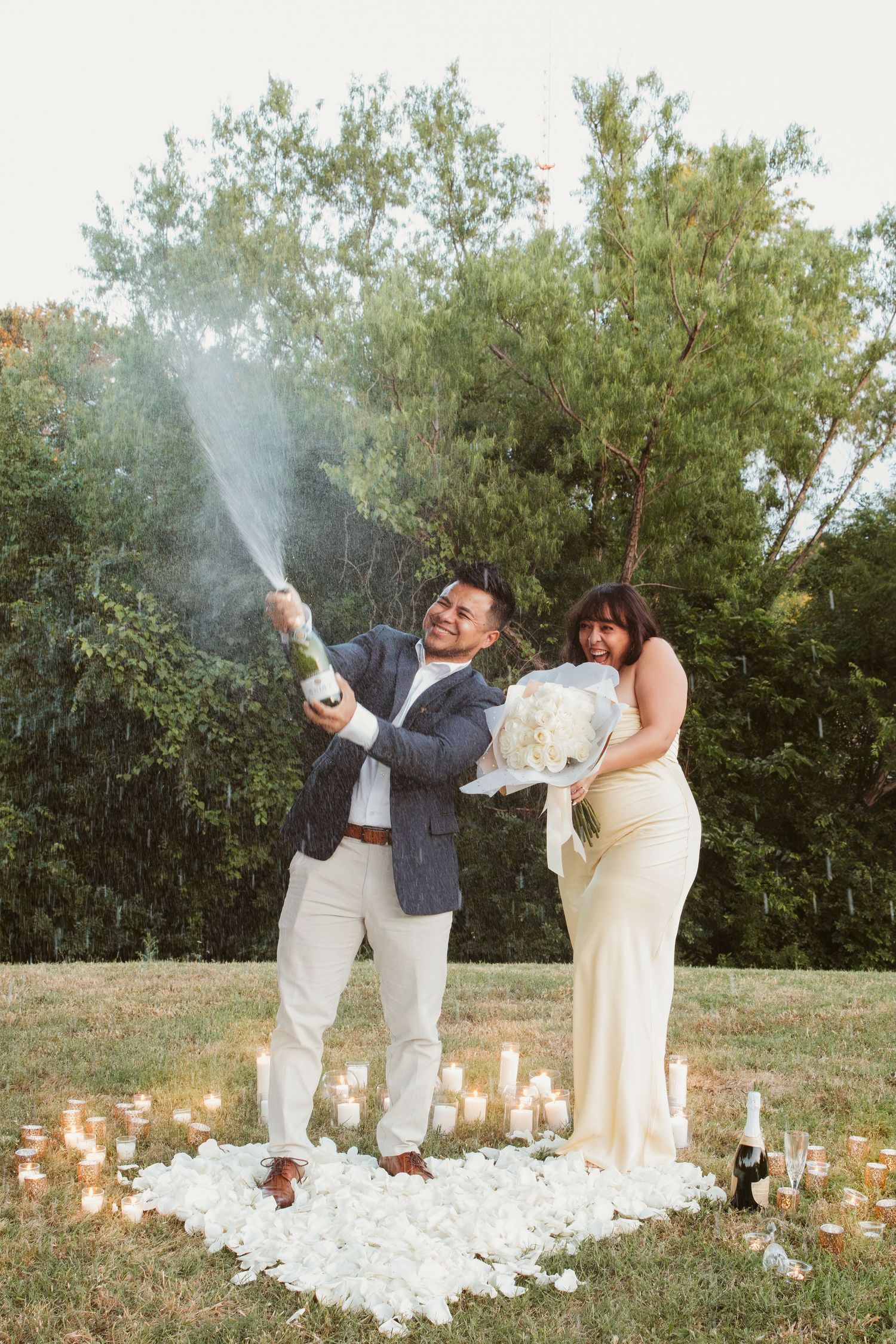 A couple celebrates on a white rug surrounded by lit candles outdoors, spraying champagne while wearing formal attire.
